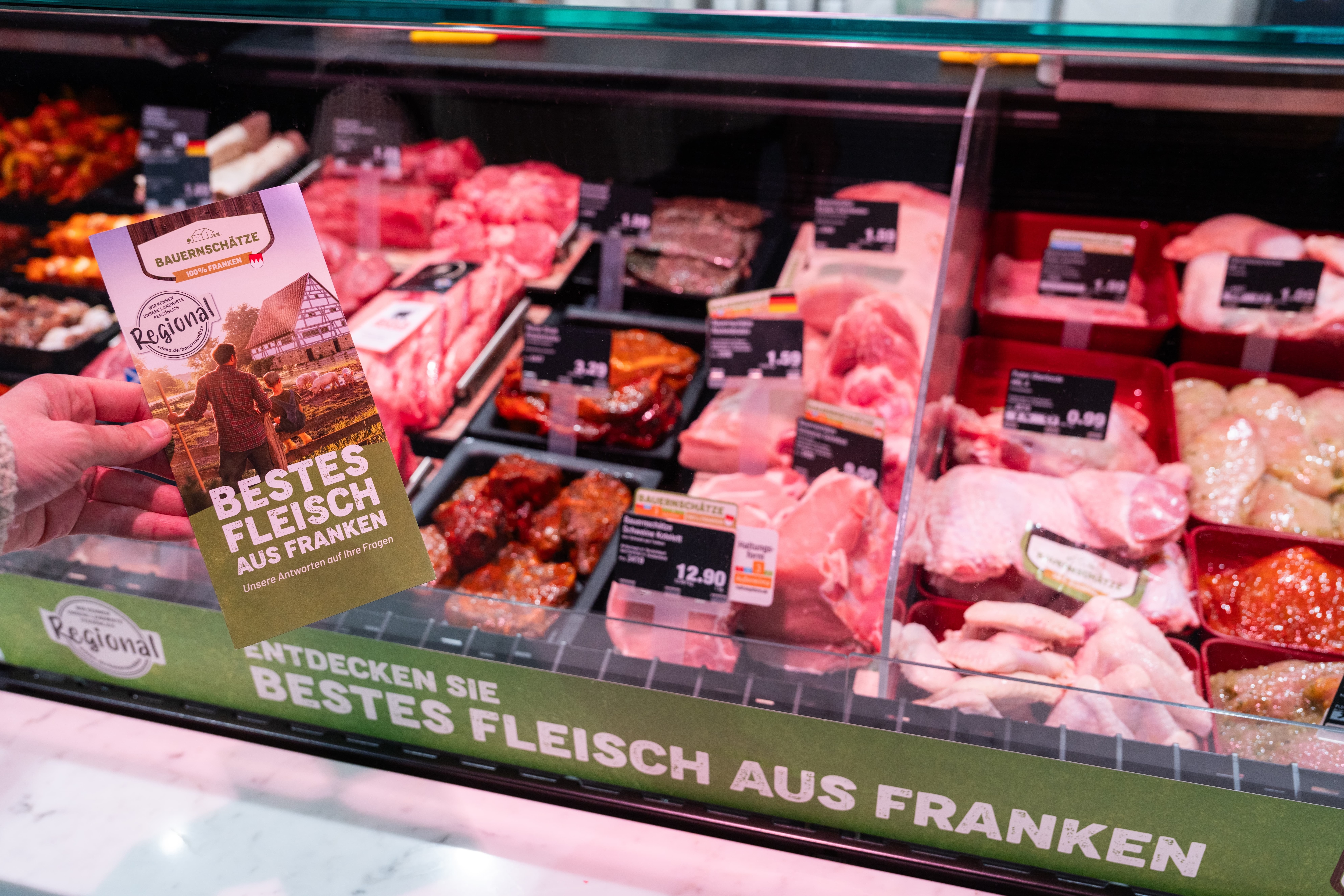Eine Hand hält einen Flyer mit der Aufschrift: "Bestes Fleisch aus Franken". Im Hintergrund eine Fleischtheke.