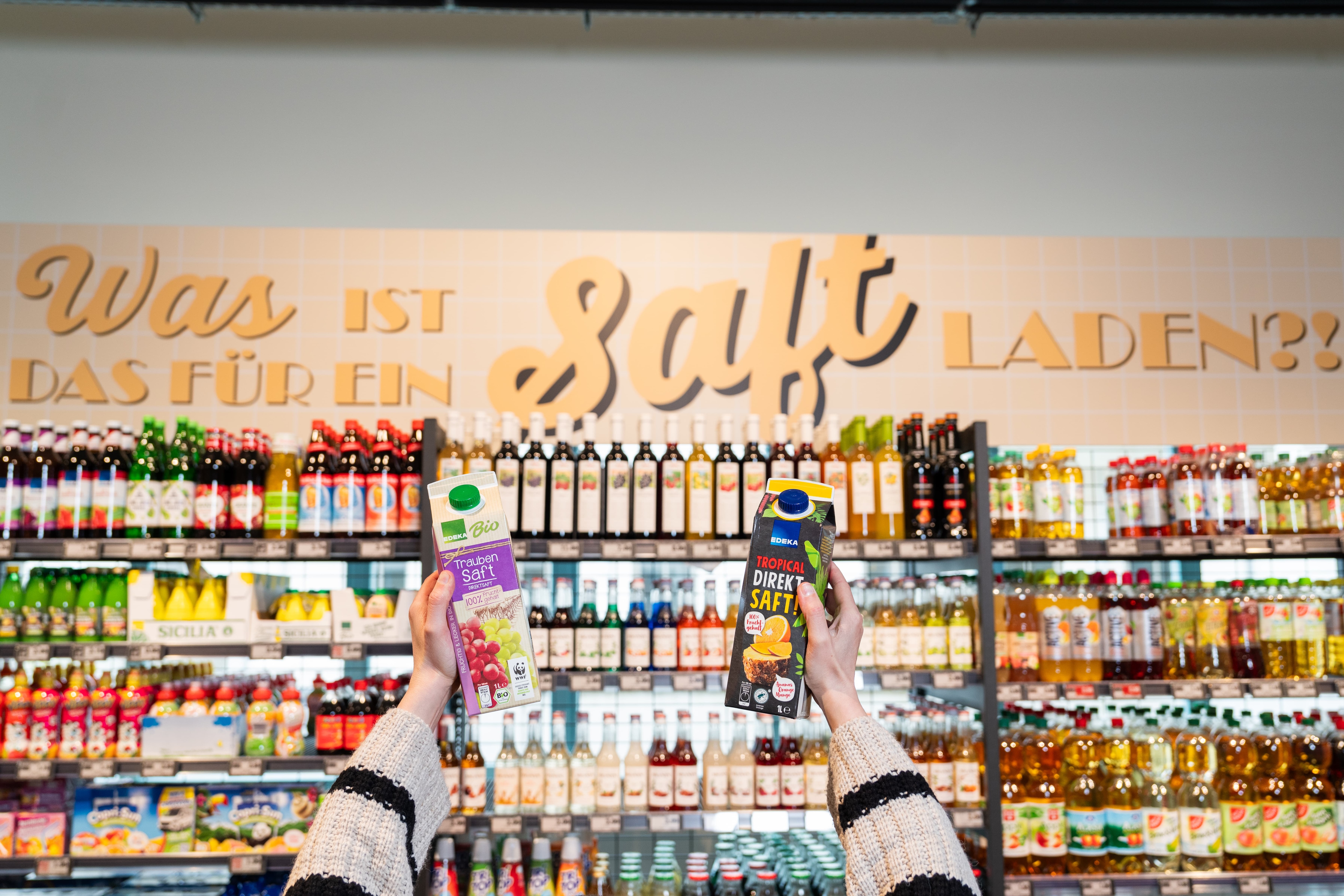 Zwei Hände halten jeweils ein Tetra Pak Saft in die Luft. Im Hintergrund sind Regale mit Säften und Softdrinks zu erkennen.