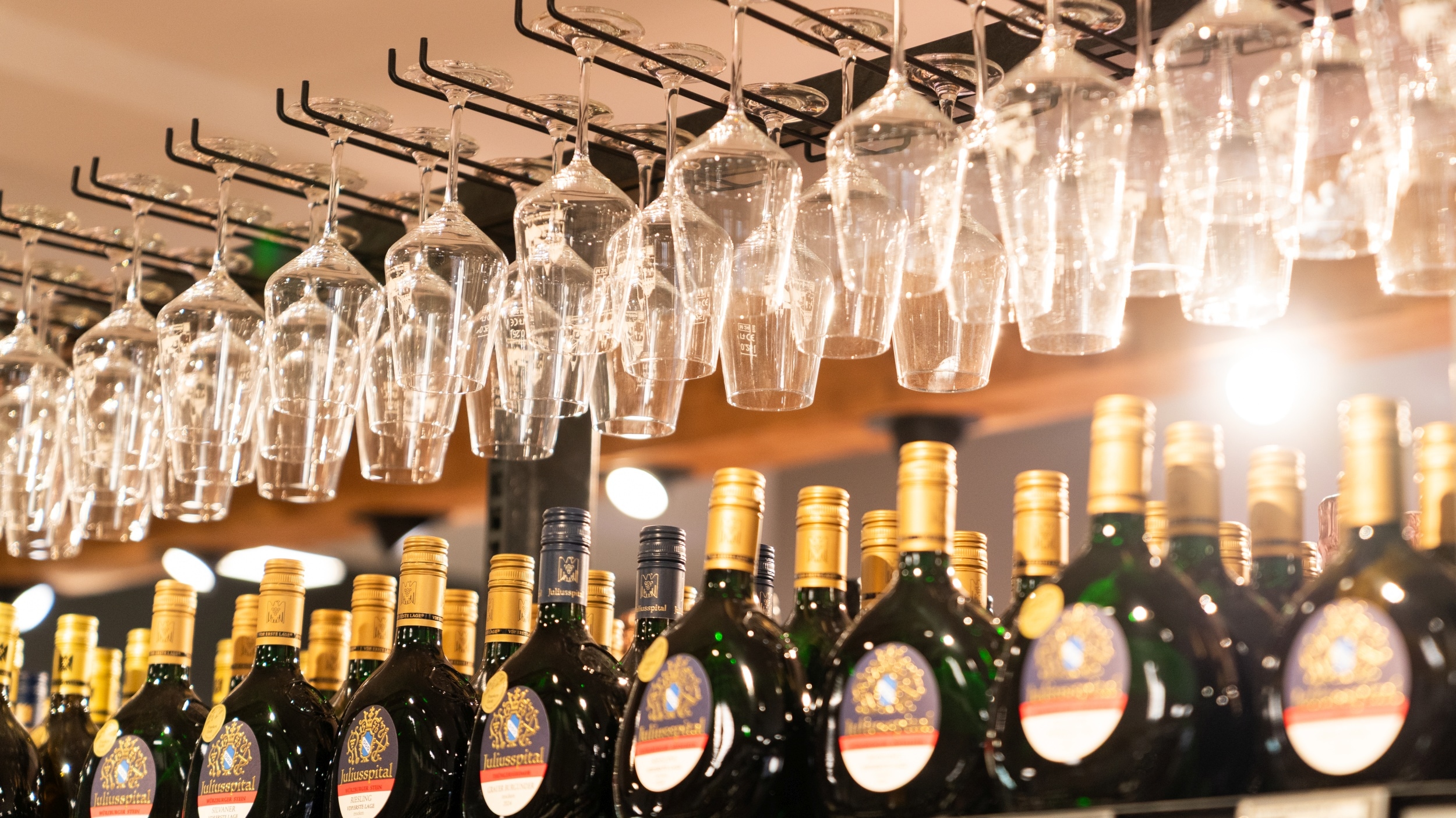 Oberer Regalausschnitt des fränkischen Weinregals. Oberhalb des Regales hängen kopfüber Weingläser, darunter fränkische Weinflaschen