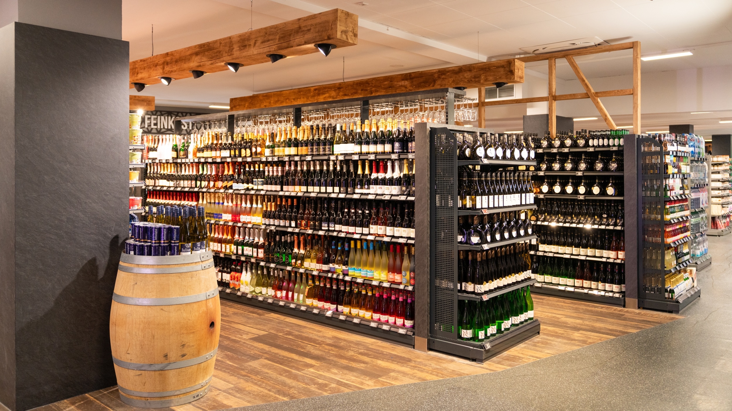 Seitliche Ansicht der Abteilung "Fränkische Weine" mit 2 Regalen mit verchiedenen Anbietern und Weinsorten. Leicht im Vordergrund ein Weinfass, auf dem Wein in Flaschen und Dosen arrangiert wurden