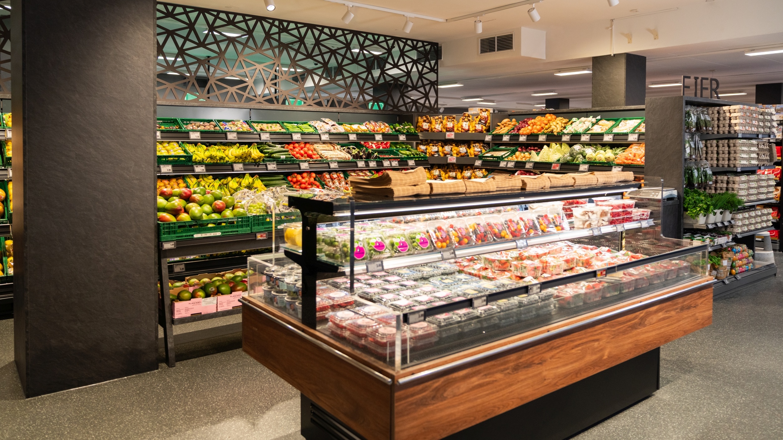 Im Vordergrund befindet sich eine SB-Theke mit portionierten und abgepackten Obst- und Salatsorten. Im Hintergrund ist ein Teil der Obst- und Gemüseabteilung zu sehen