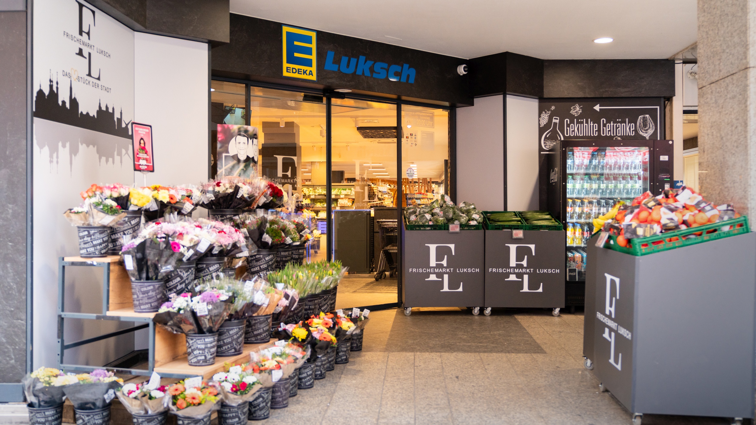 Eingangsbereich des Edeka-Marktes mit Blumenstand auf der linken Seite, Angeboten von Obst- und Gemüse auf der rechten Seite und der Eingangstür und Einem Bereich mit gekühlten Getränken