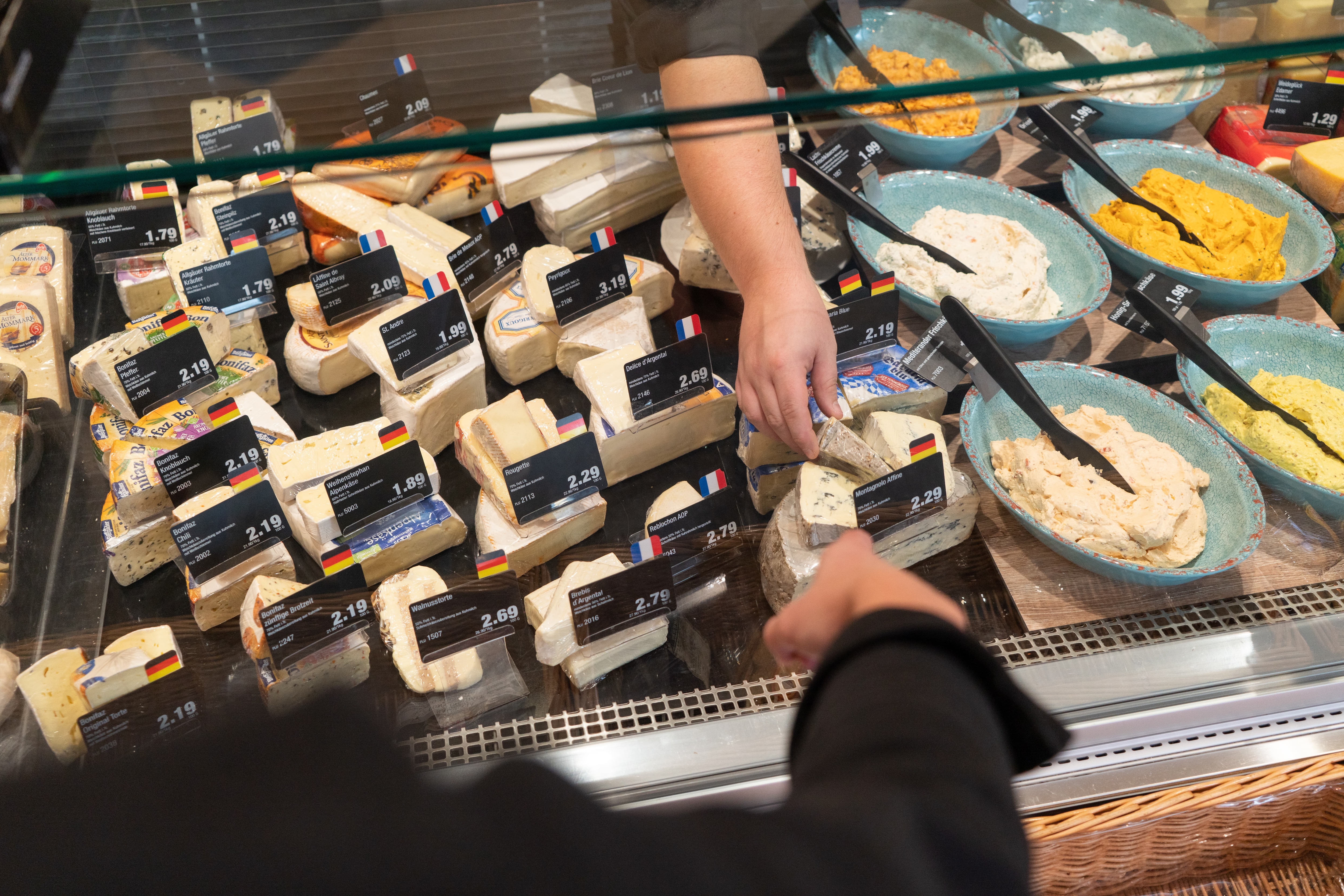 Eine Hand nimmt ein Stück Käse aus der Auslage einer Frischetheke.