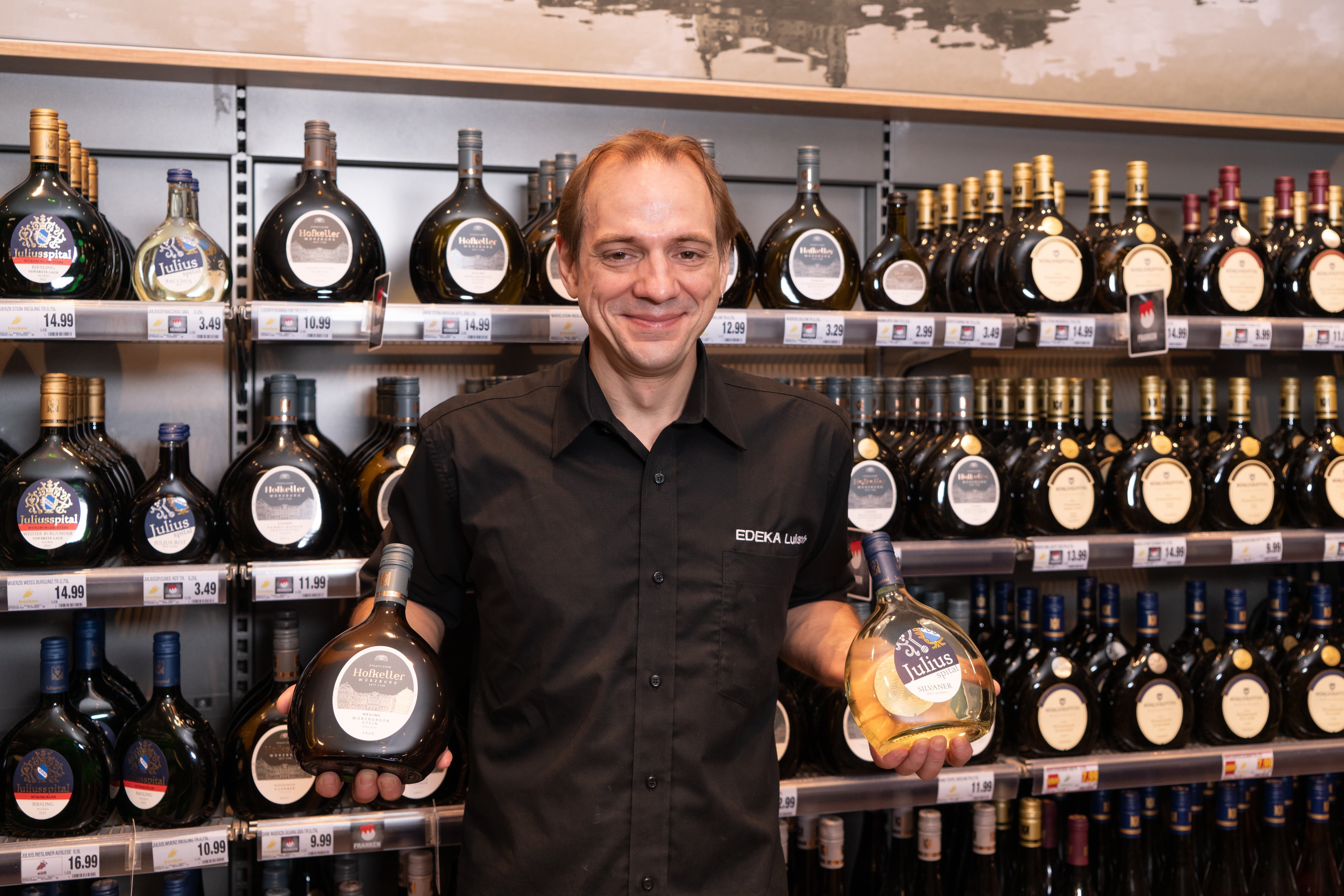 Ein lächelnder Mitarbeiter hält in jeder Hand jeweils eine Flasche Wein.