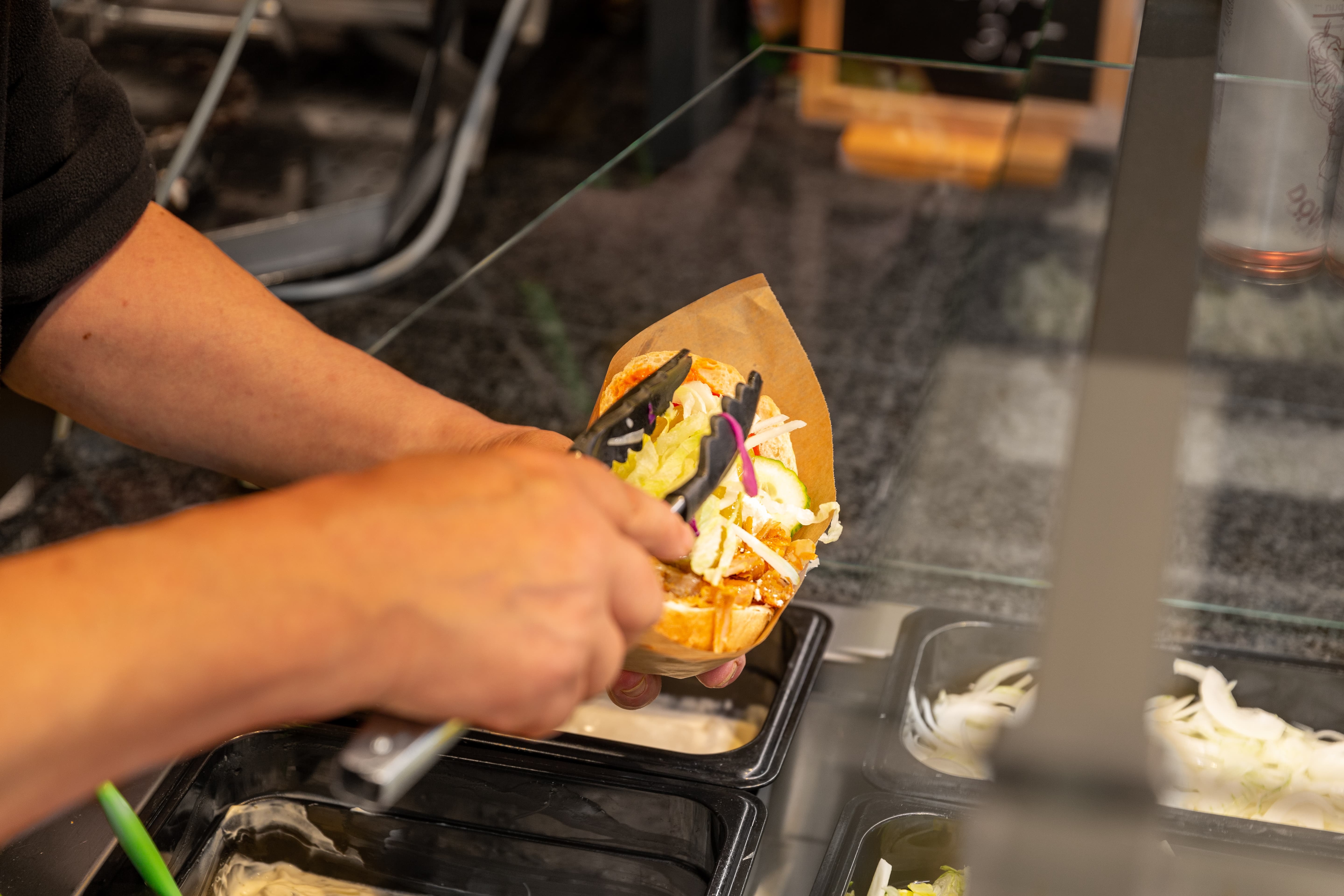 Eine Hand hält ein Döner während die andere weitere Zutaten mit einer Zange in den Döner füllt.
