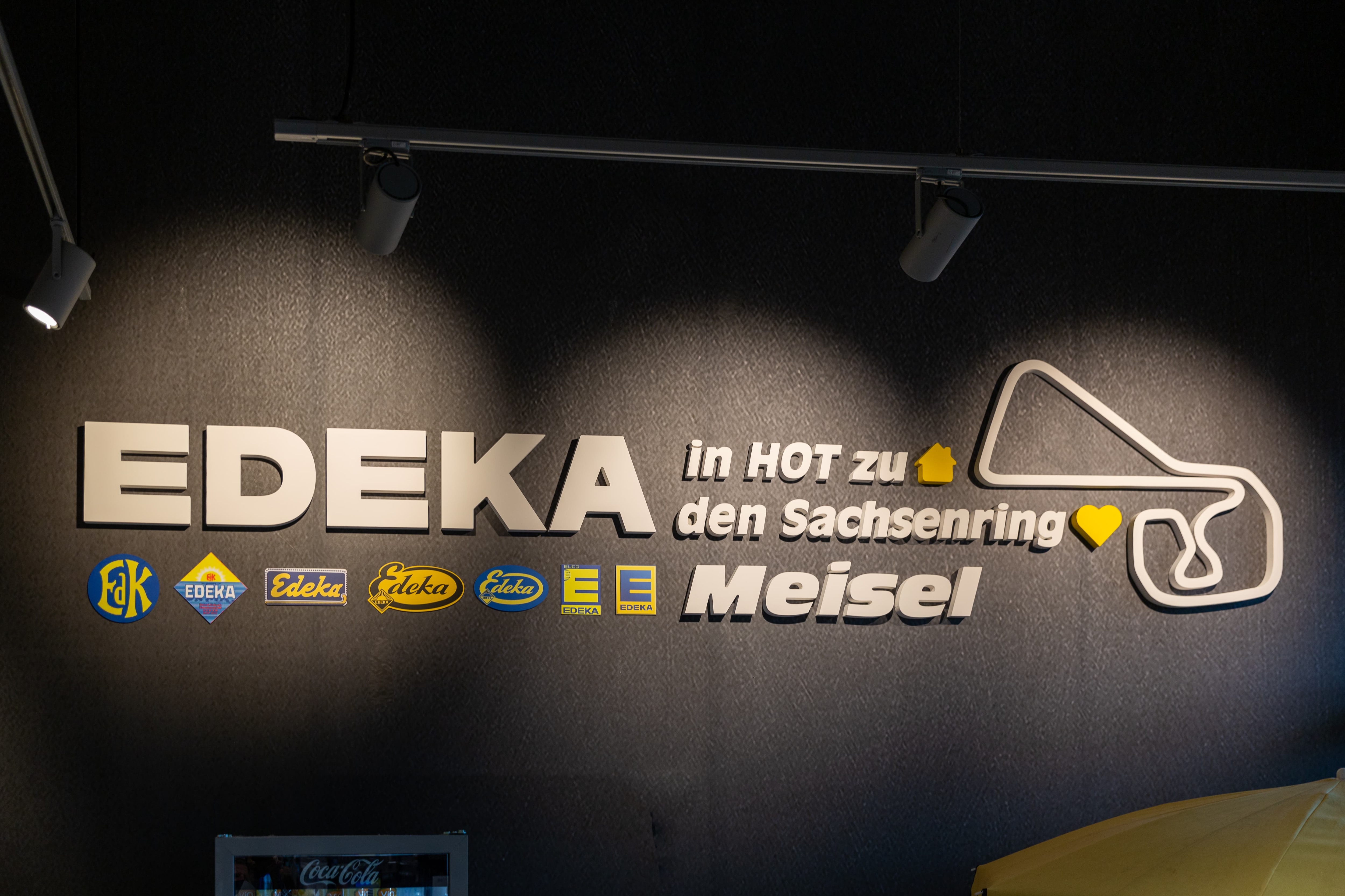 Ein weißer Edeka-Schriftzug mit regionalem Bezug auf schwarzem Grund. Darunter die verschiedenen historischen Edeka-Logos.