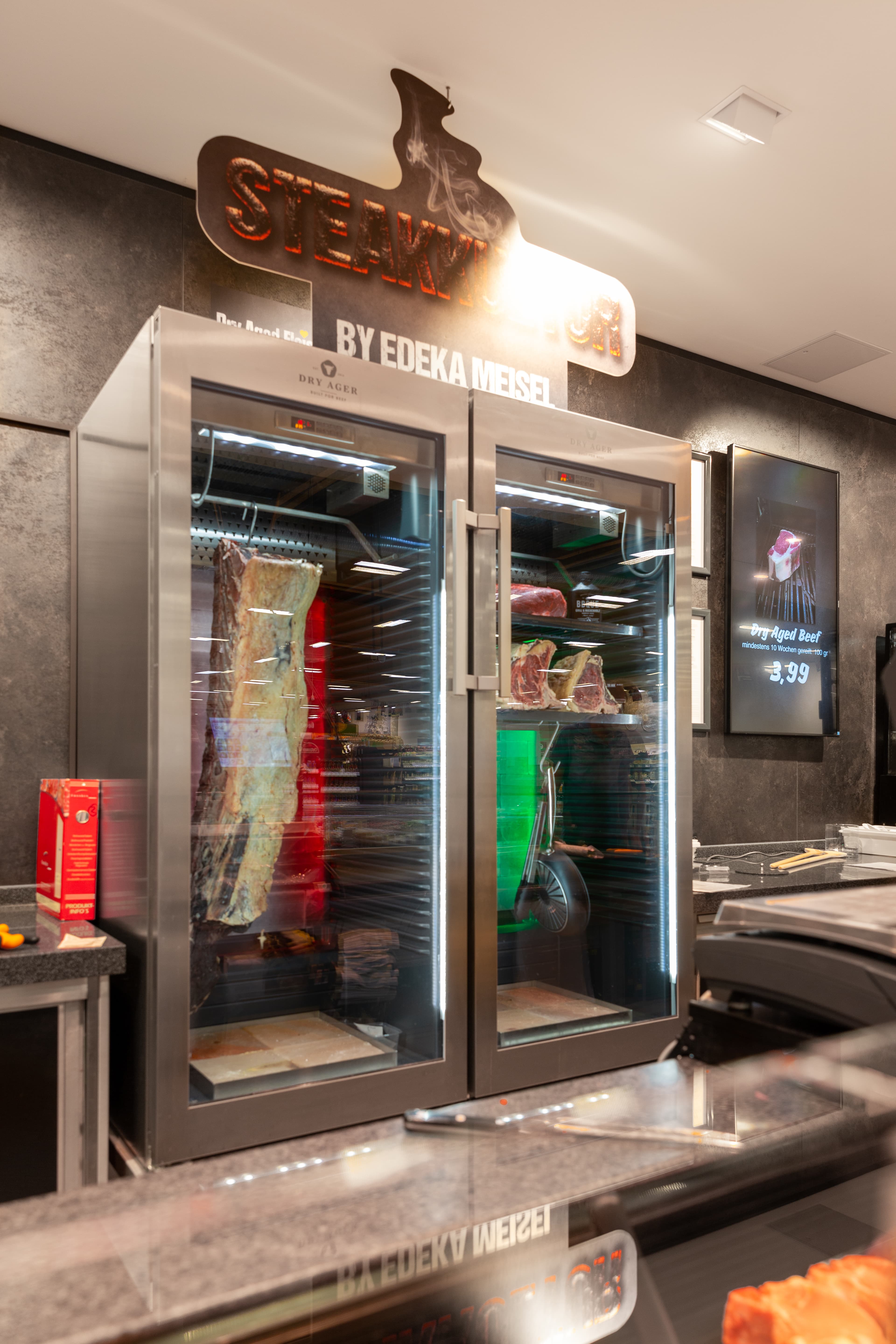 Ein Schrank mit zwei Glastüren in dem Fleisch lagert. Auf der linken Seite hängt ein großes Stück Fleisch. Auf der rechten Seite lagern kleinere Stücke Fleisch auf Blechen.