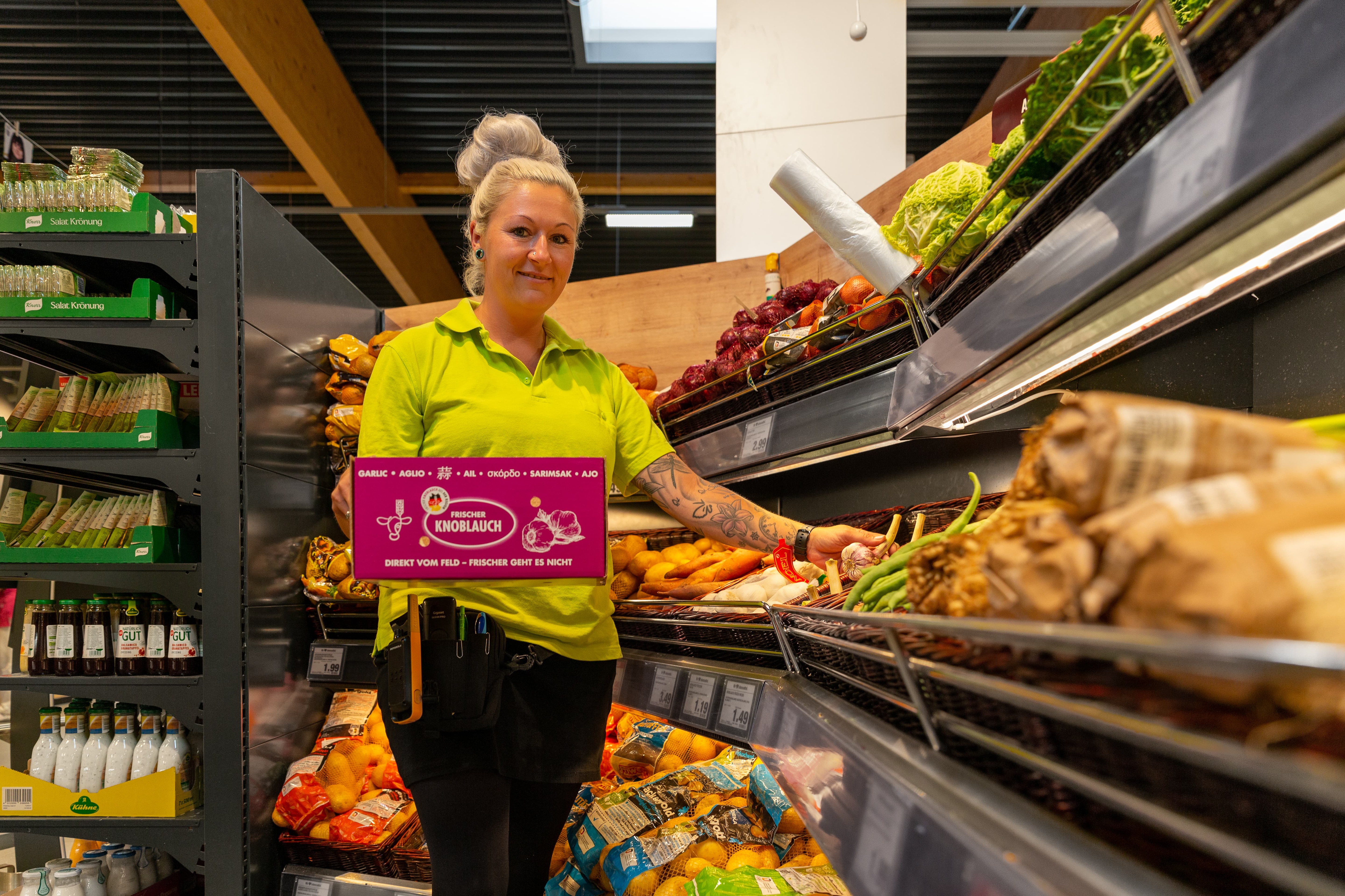 Eine Edeka-Mitarbeiterin hält eine Kiste Knoblauch in der einen Hand. Mit der anderen verräumt sie den Knoblauch.