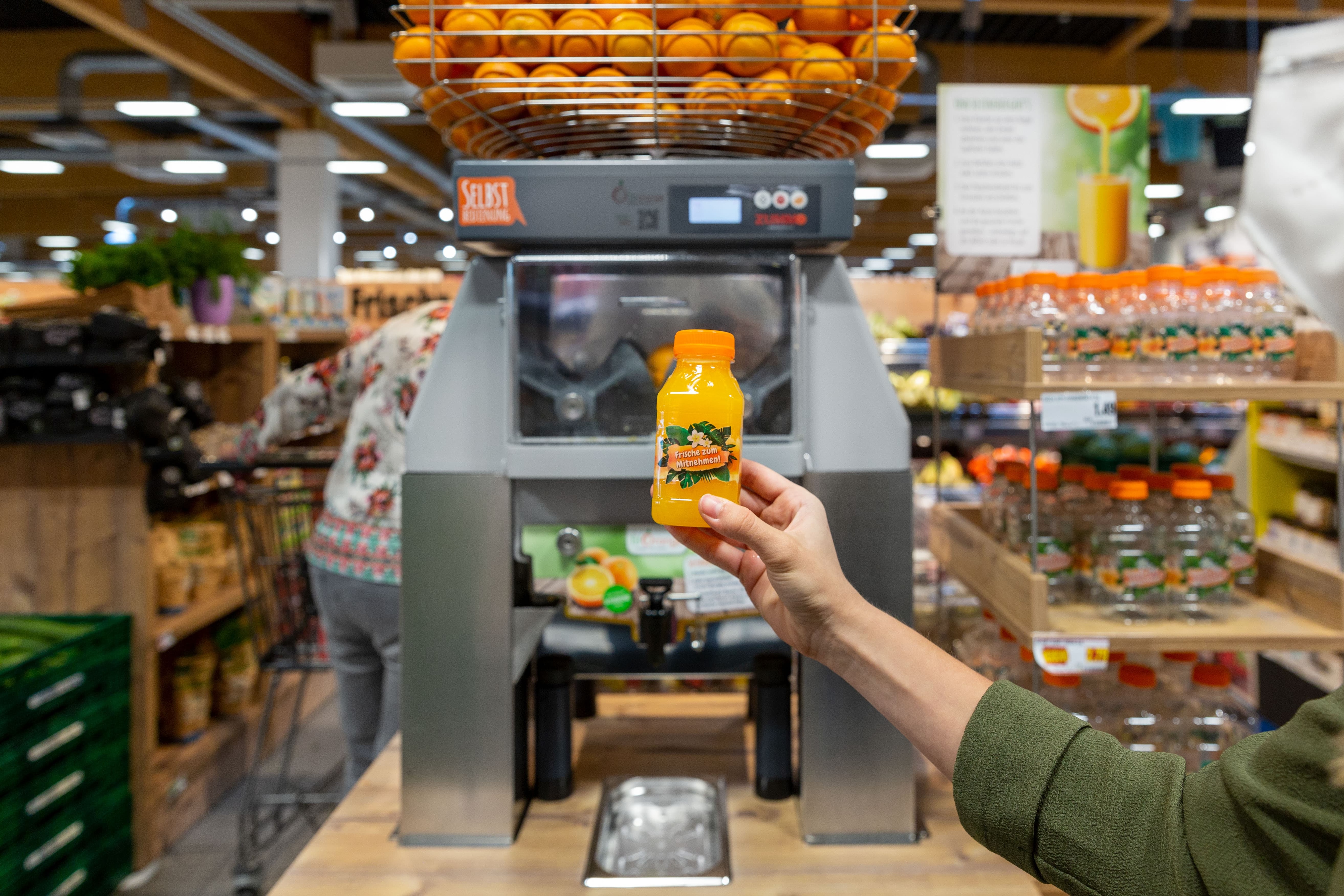 Im Vordergrund eine Hand die eine Flasche frisch gepressten Orangensaft hält. Im Hintergrund eine Saftpresse bestückt mit Orangen.