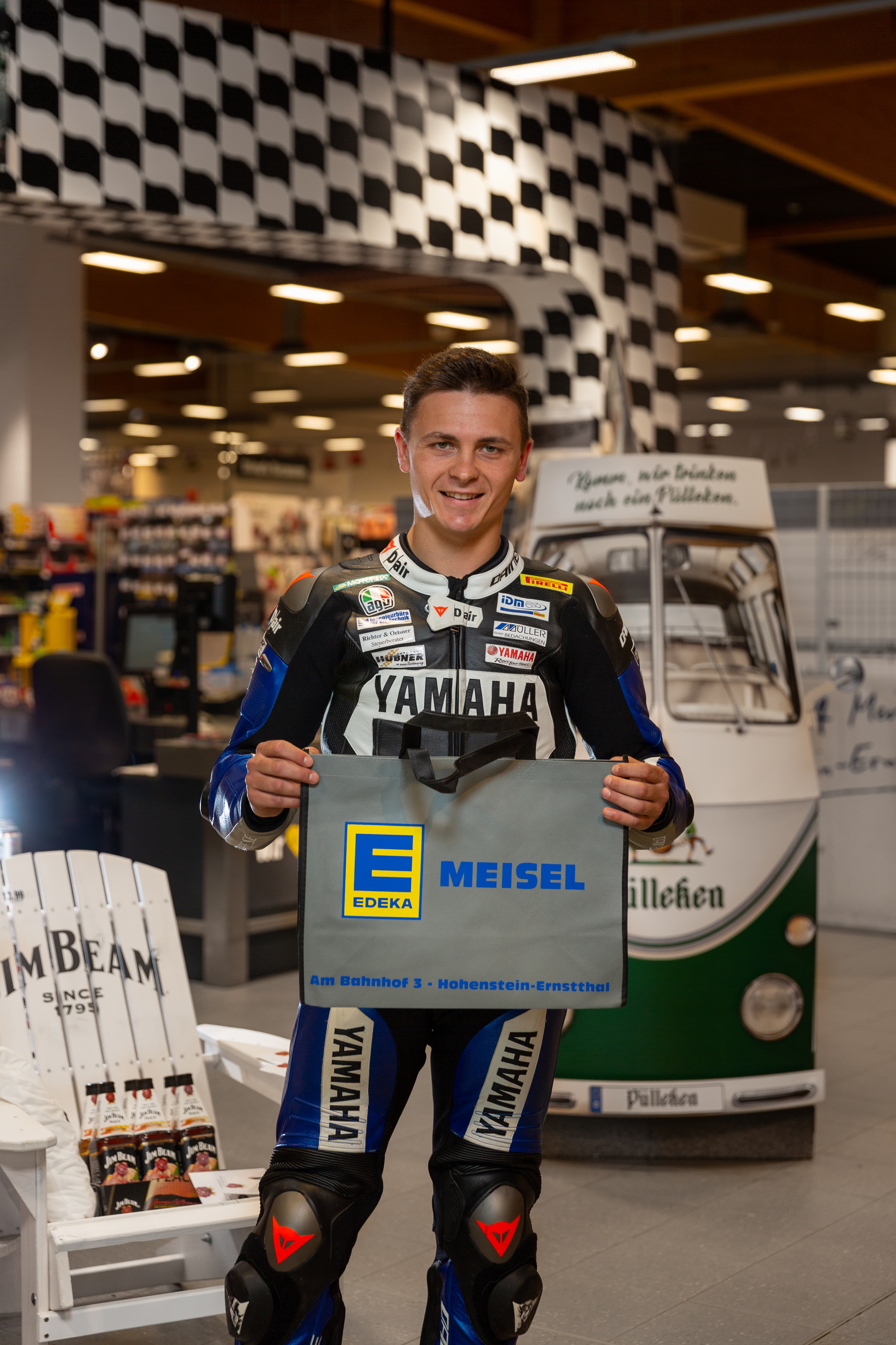 Ein Mann in einem Rennanzug hält einen Mehrwegbeutel eines Edeka-Marktes in die Kamera.