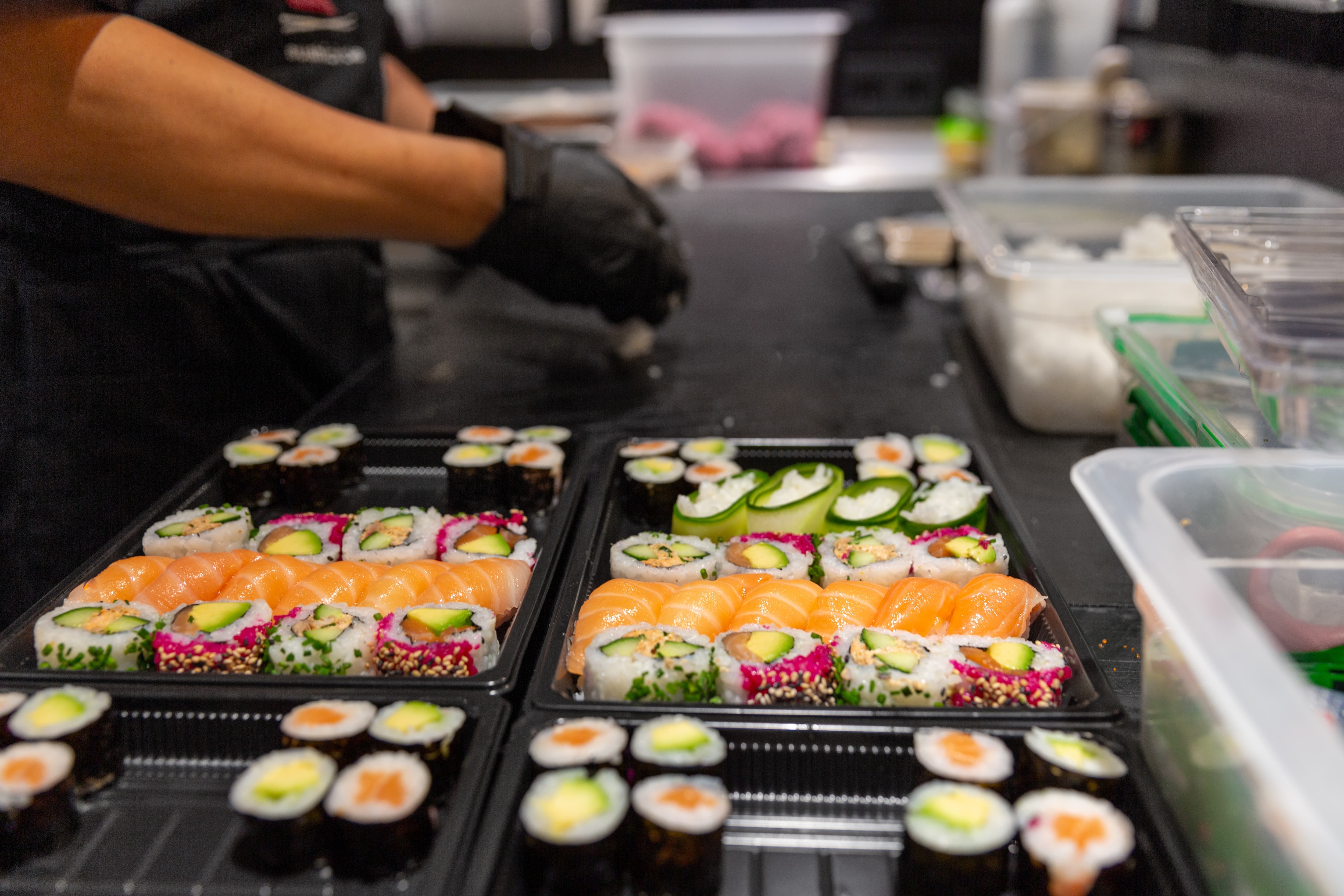 Im Vordergrund frisch zubereitetes Sushi. Im Hintergrund rollt eine Person Sushi.