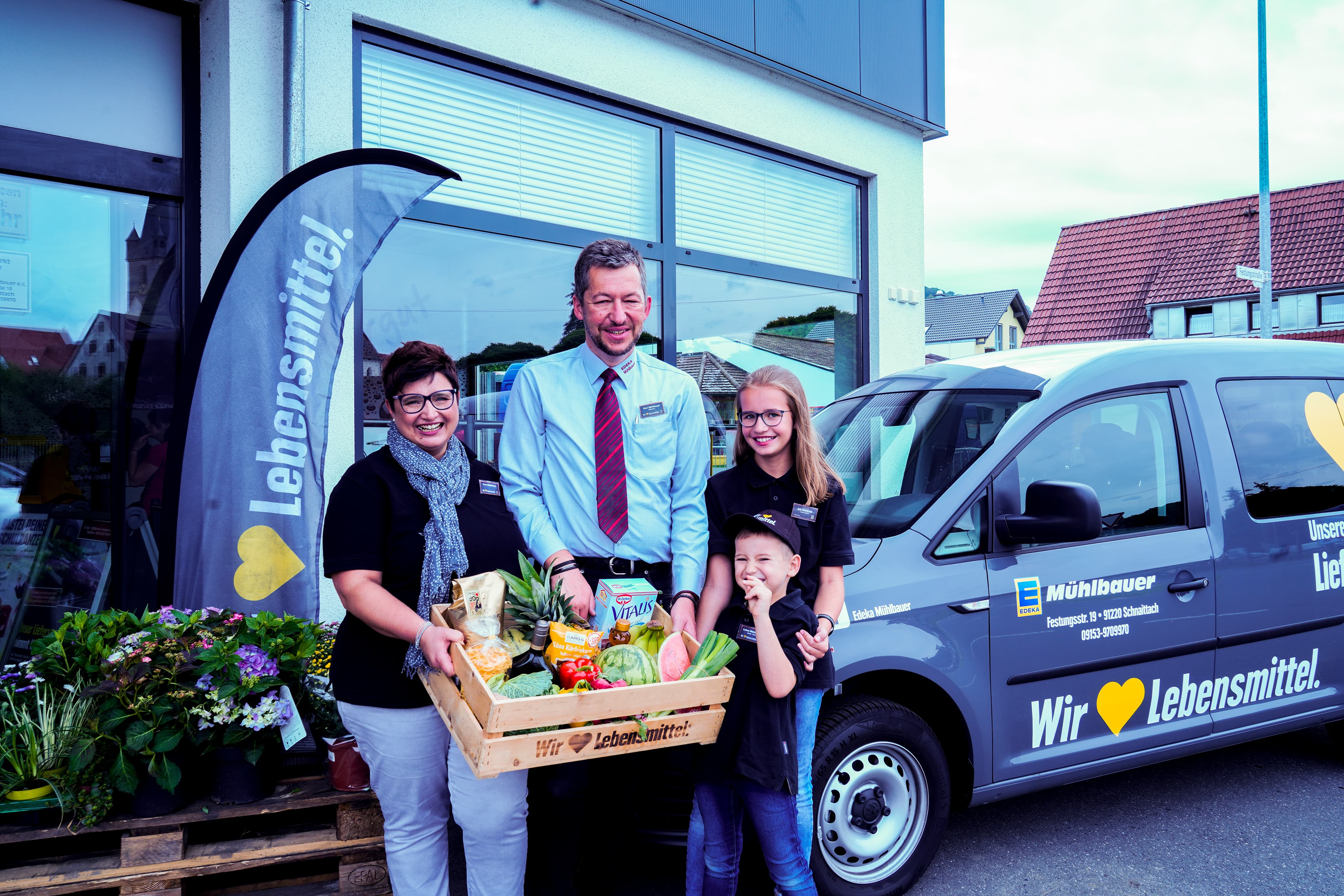 Die Inhaberfamilie vor dem Markt. Gemeinsam halten sie eine Holzkiste mit Lebensmitteln hoch.