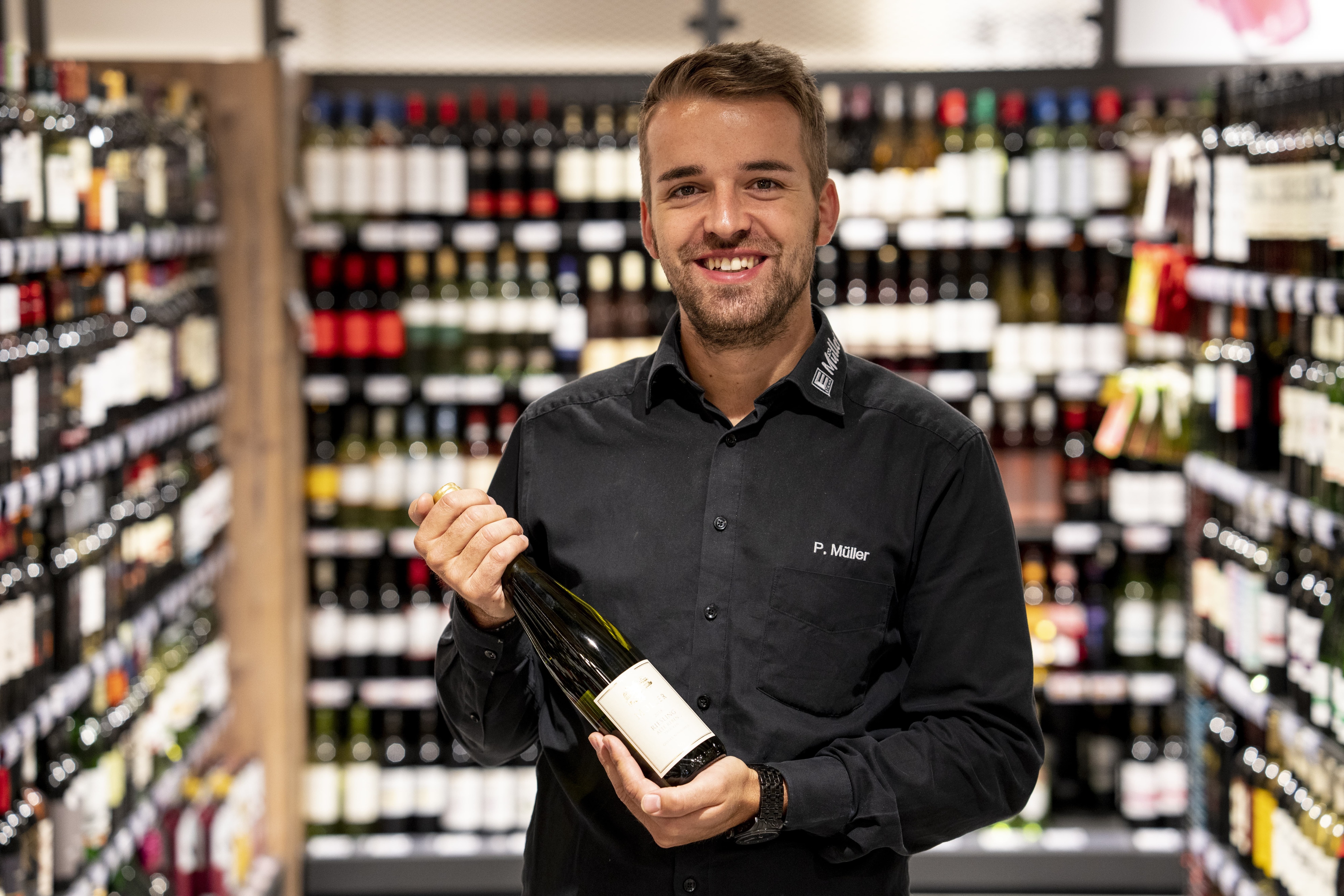 Der Marktleiter, lächelnd mit einer Flasche Wein in der Hand, im Hintergrund verschwommen die Weinabteilung.