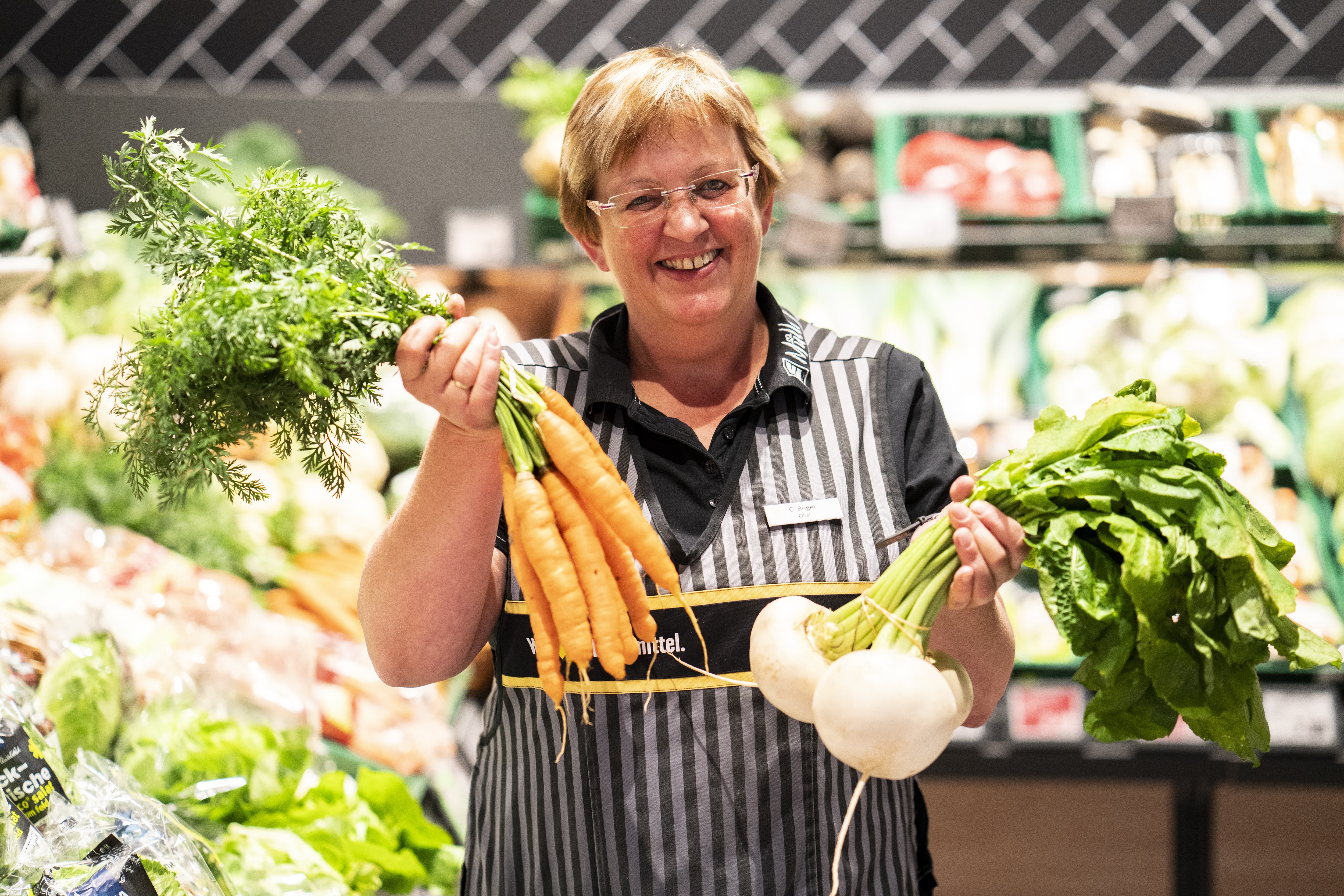 Eine Mitarbeiterin in der Obst- und Gemüseabteilung hält Gemüse hoch. Sie lächelt.