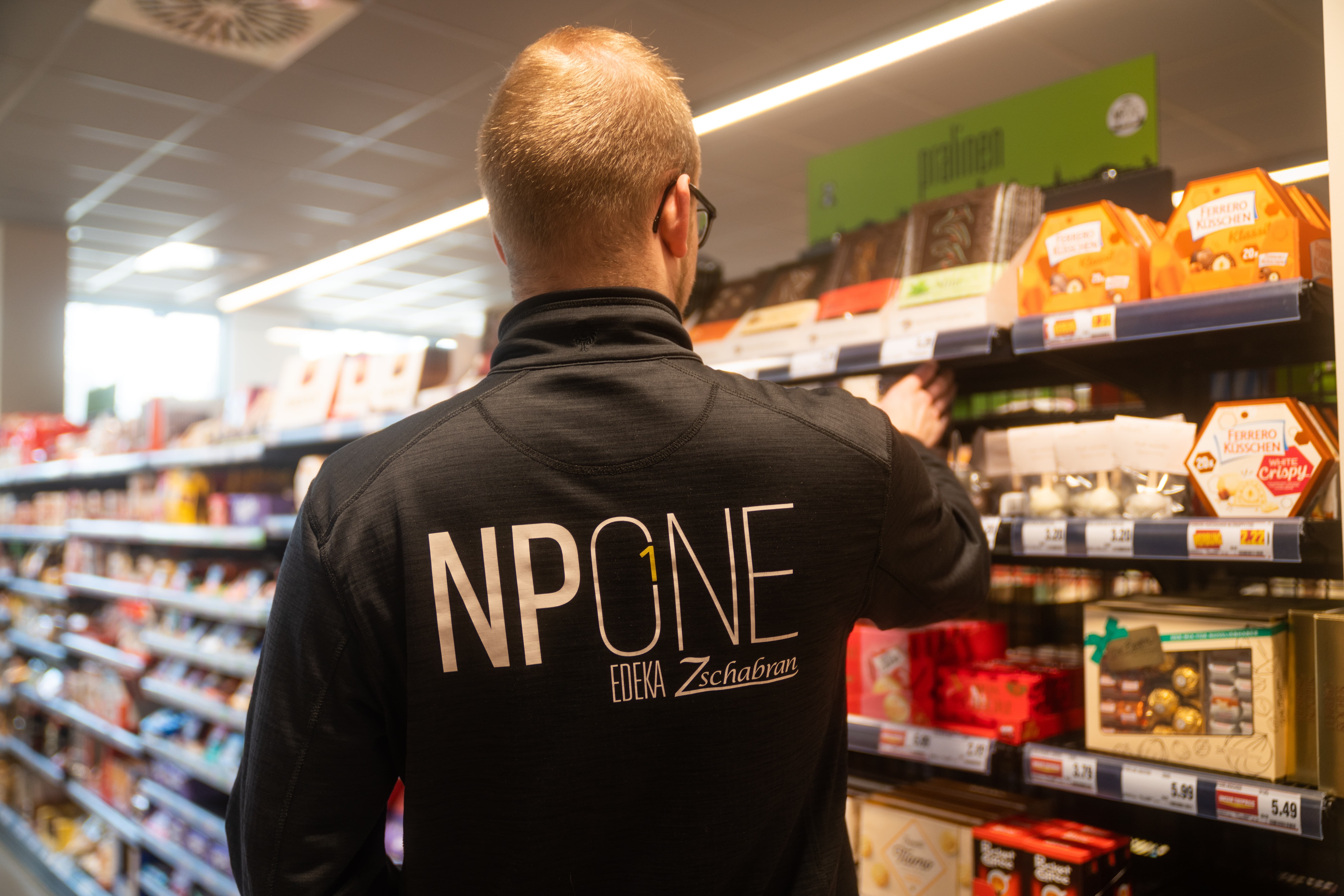 Ein Mann von hinten am Pralinenregal. Auf der Rückseite seiner Jacke steht NPONE EDEKA Zschabran.