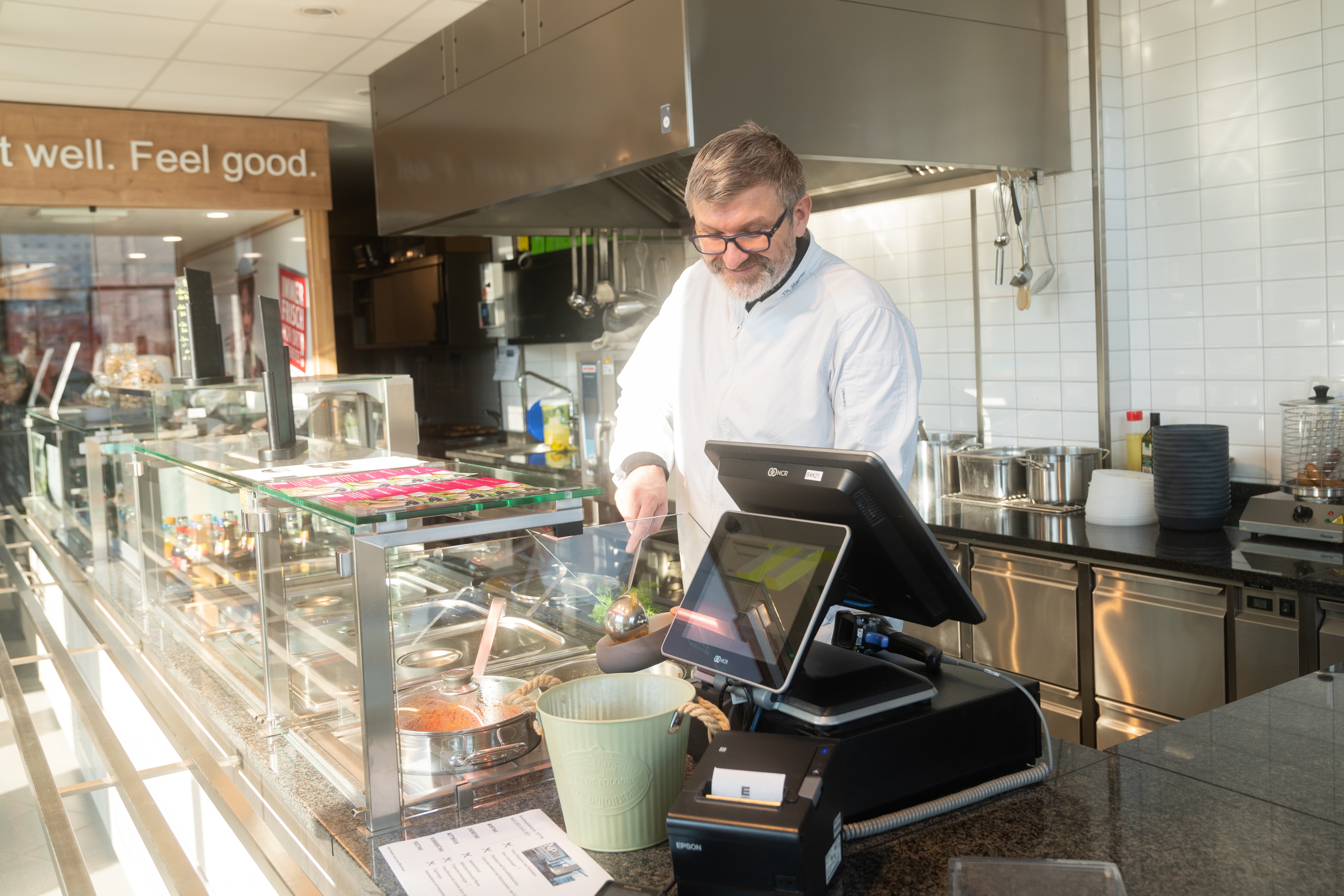 Hinter einem Tresen füllt ein Mann in weißem Kittel eine Schale mit Essen auf.