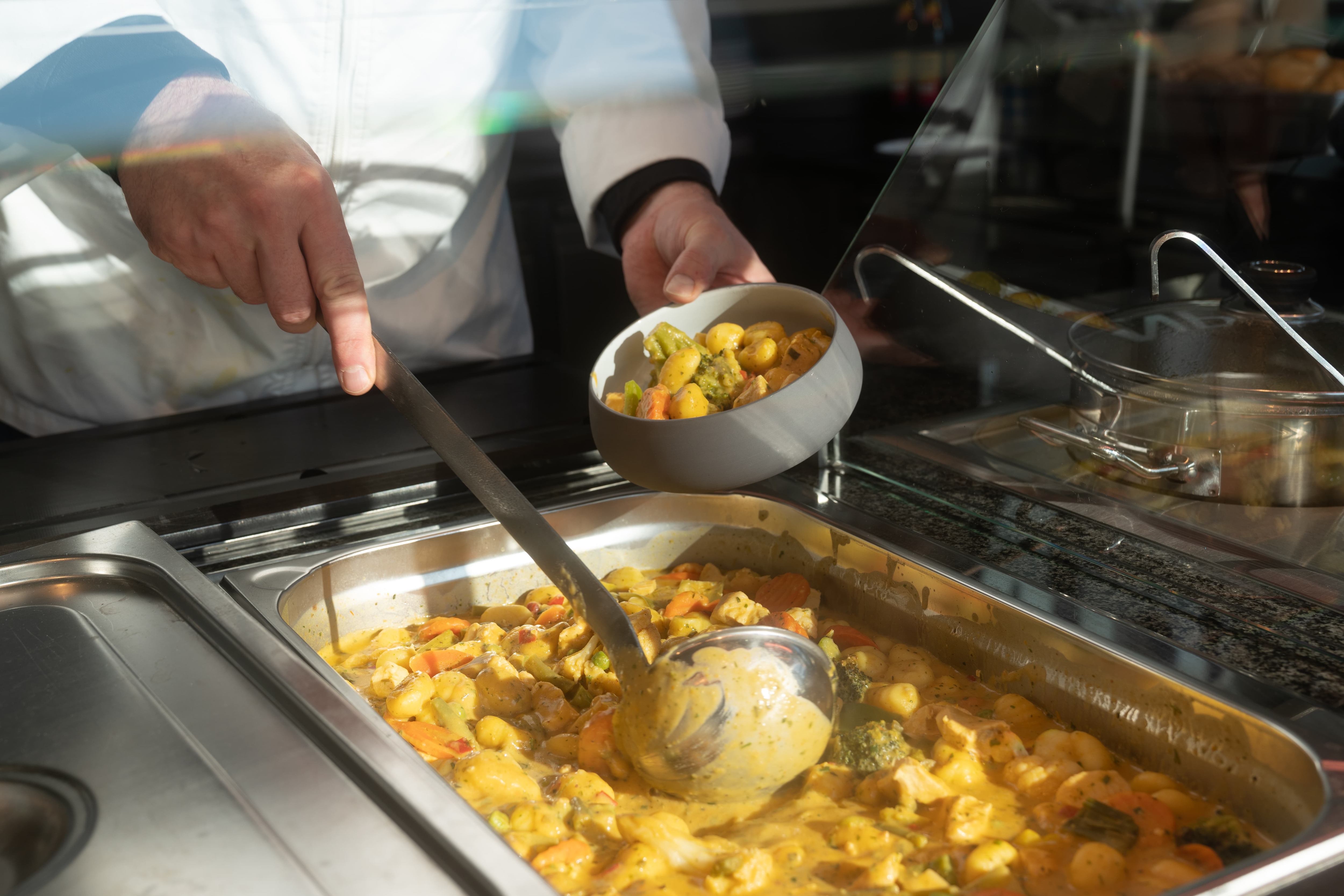 Mann mit weißem Kittel füllt eine Portion Gemüseauflauf in ein Schälchen.