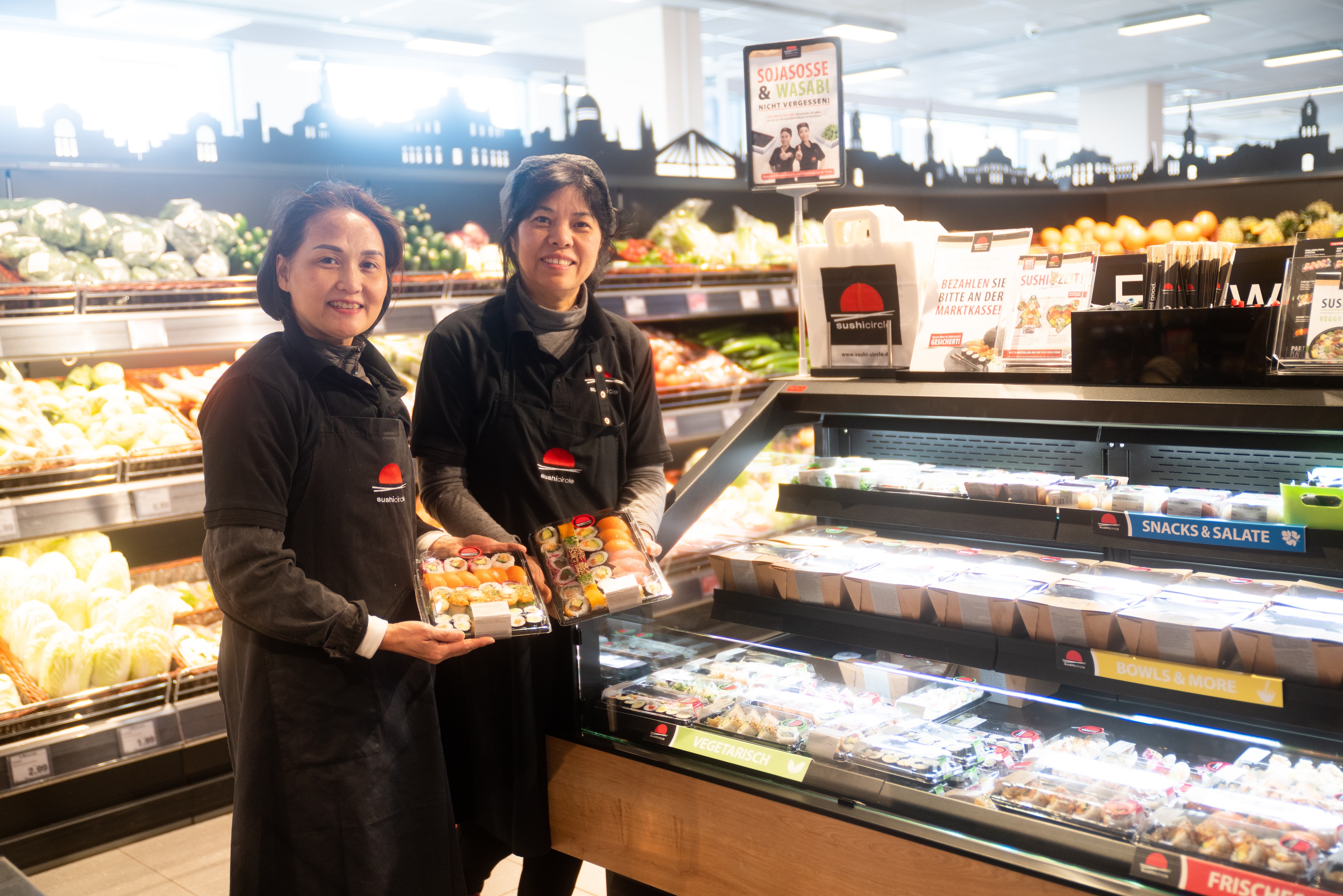 Zwei Mitarbeiterinnen mit schwarzen Schürzen halten jeweils eine Packung Sushi in der Hand.