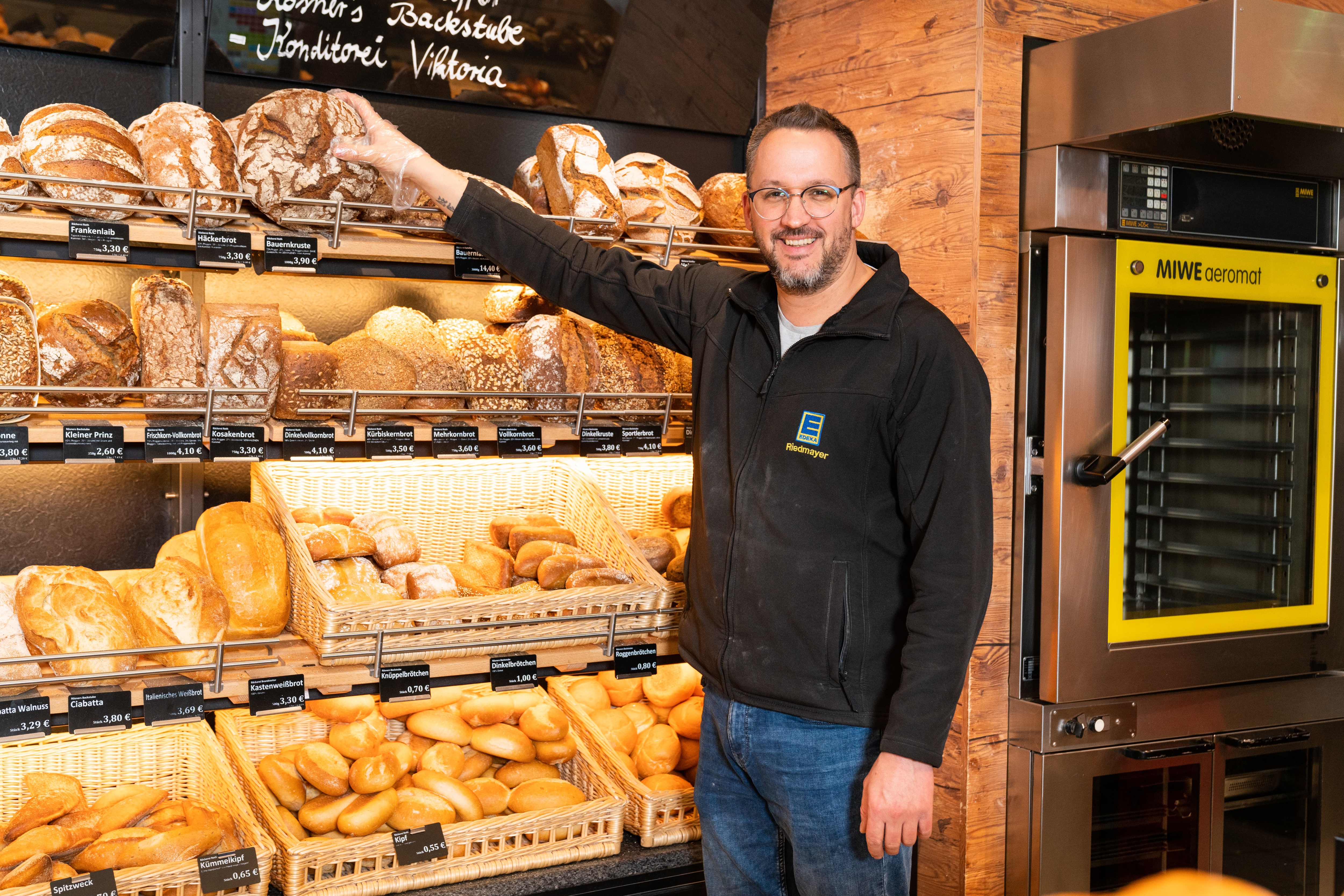 Ein Mann in schwarzer EDEKA-Jacke greift mit einem Einweghandschuh nach einem runden, braunen Brot.