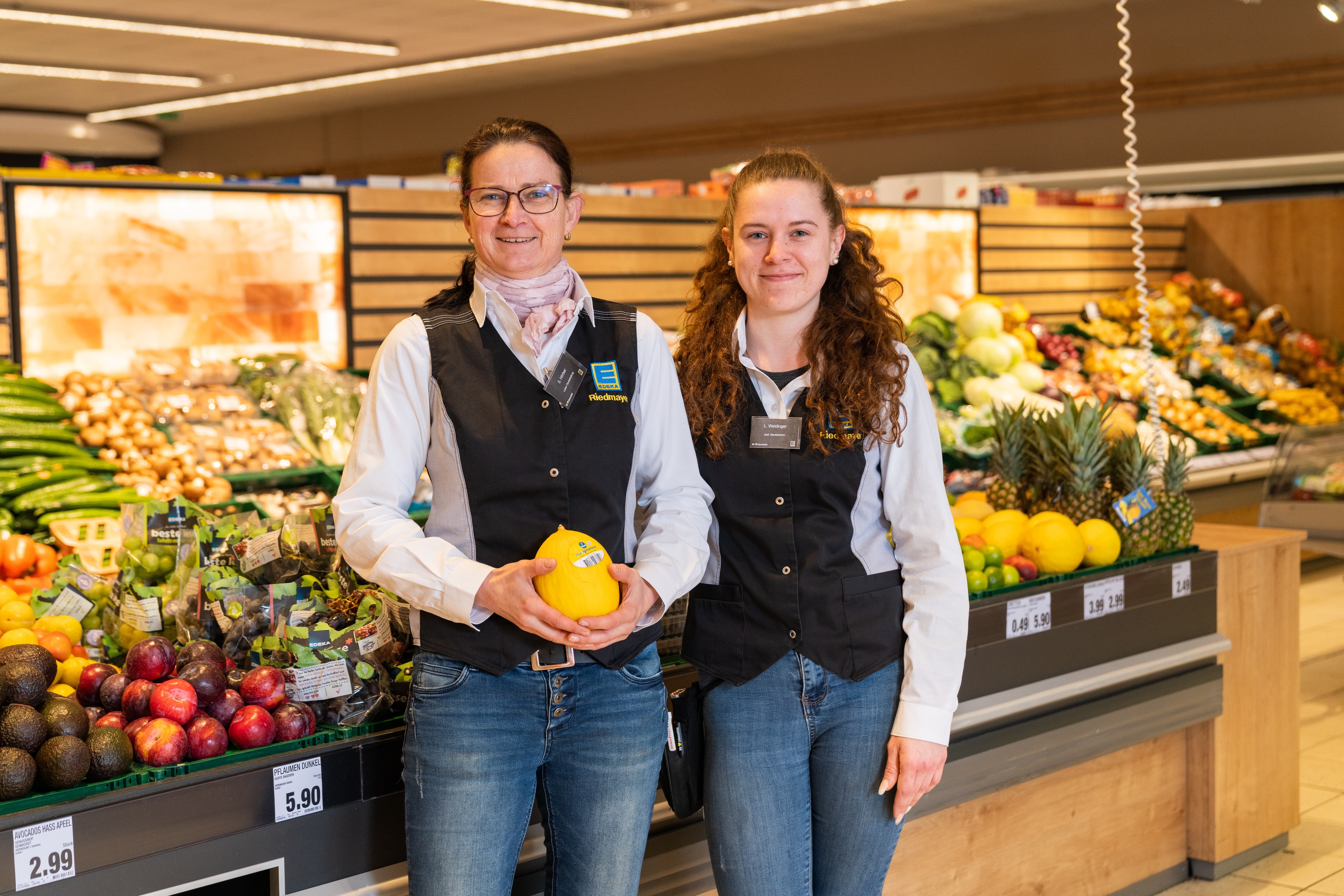 Zwei Frauen in EDEKA-Kleidung stehen lächelnd vor dem Obstregal. Eine hält eine kleine Honigmelone in der Hand.
