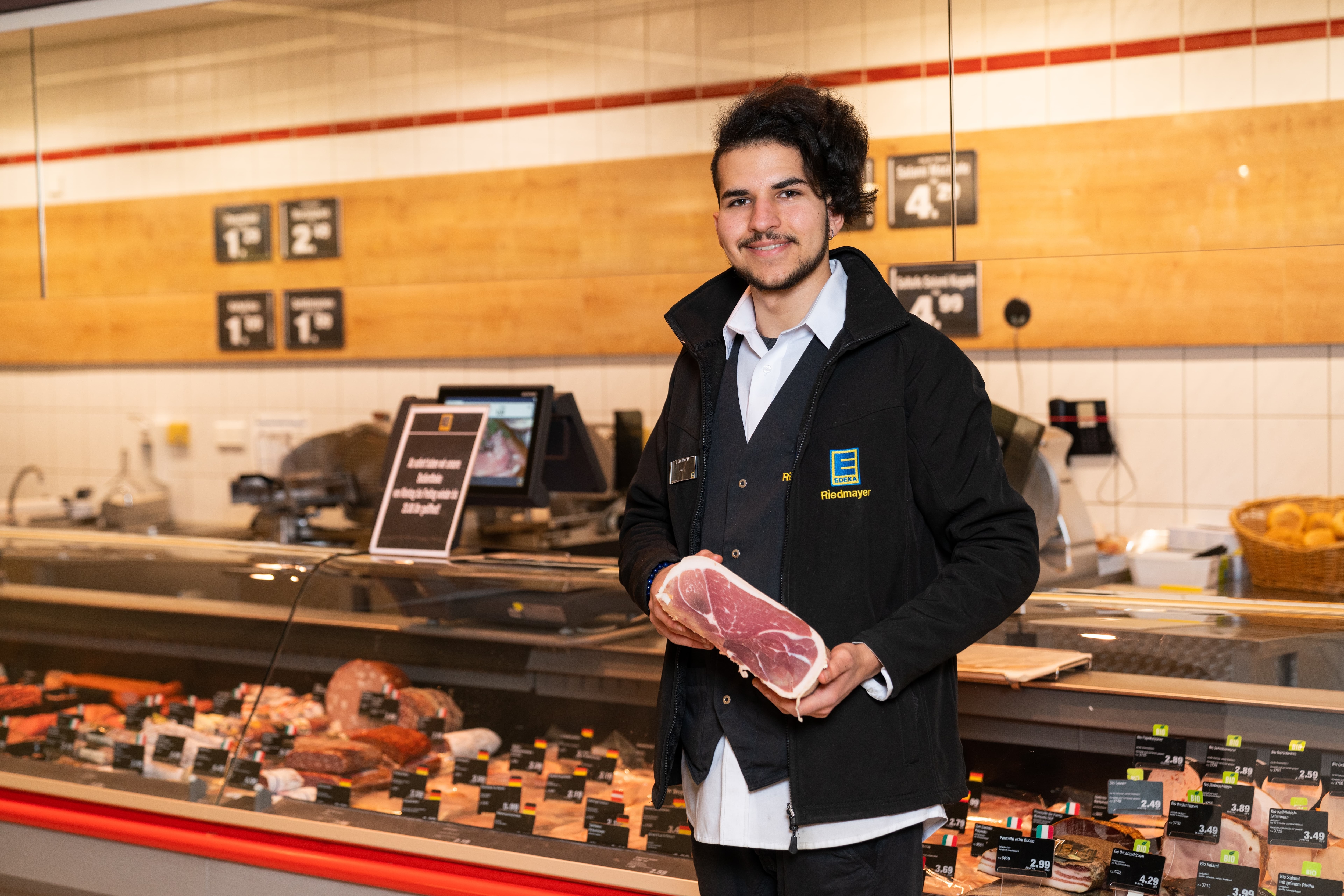 Ein junger Mann mit langen dunklen Haaren steht vor der Frischetheke und hält einen rohen Schinken vor sich. Er trägt eine schwarze EDEKA-Jacke.