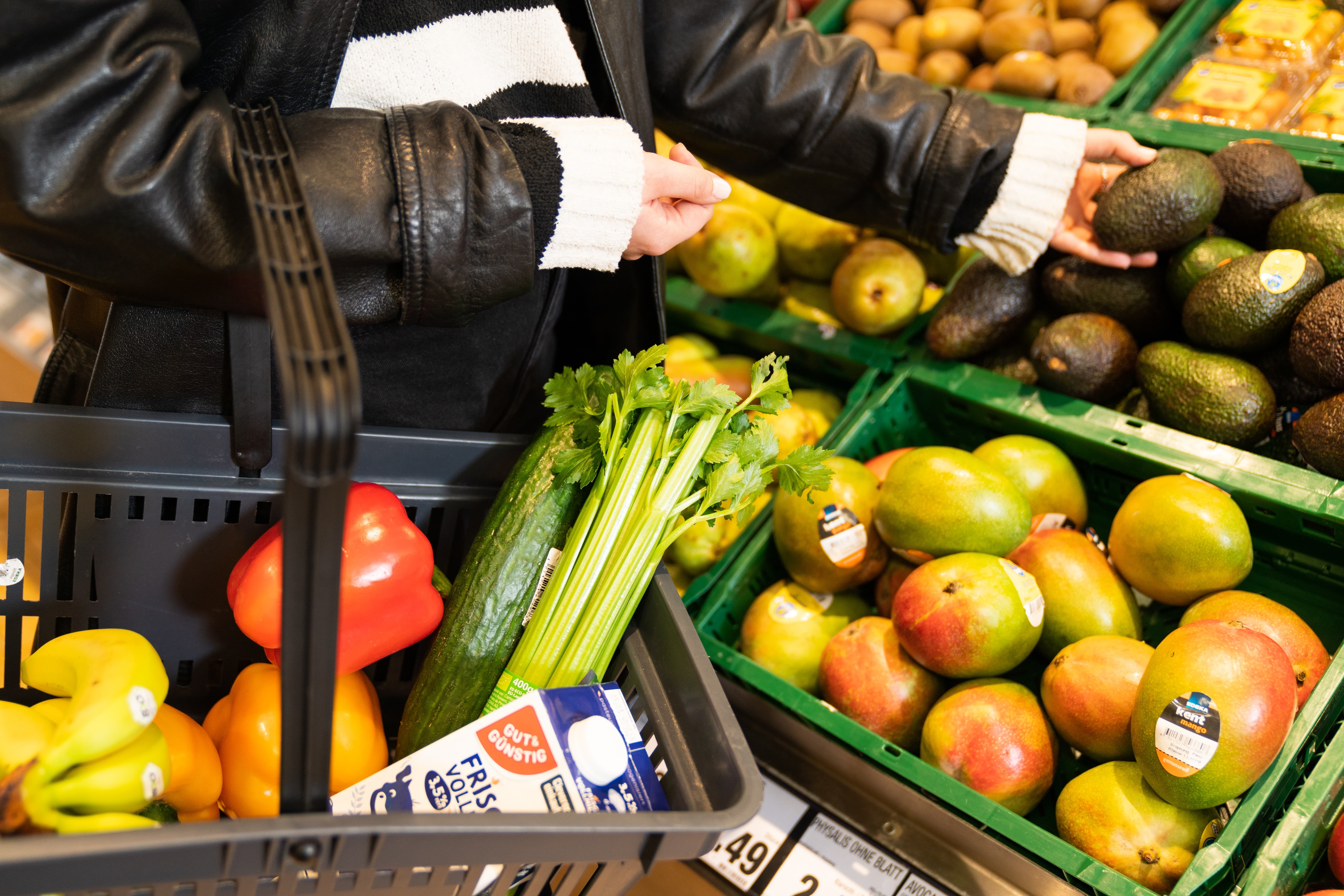Eine Frau mit Einkaufskorb betrachtet eine Avocado. Im Korb sind Bananen, Paprika, Staudensellerie, eine Gurke und Milch.