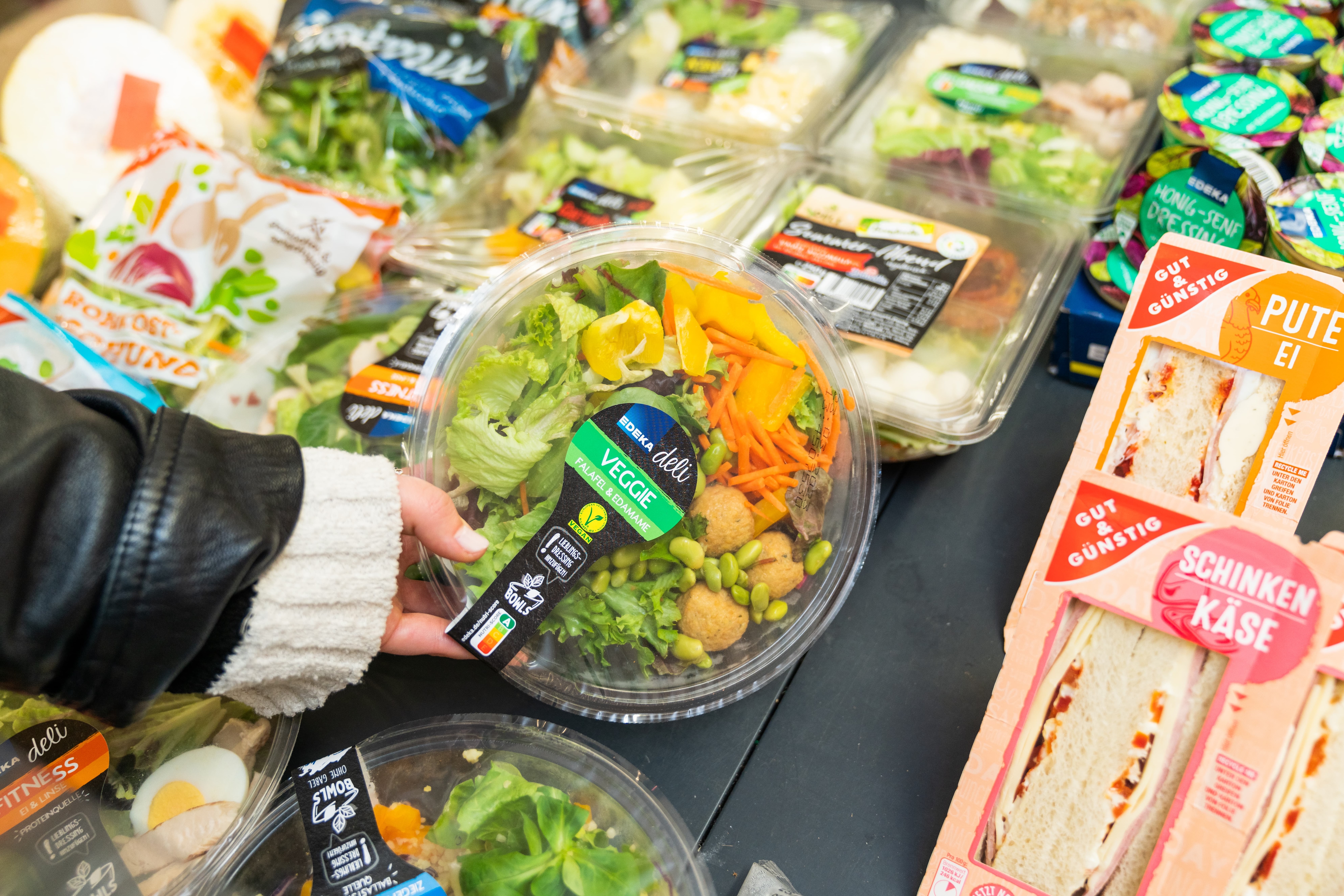 Eine weibliche Hand hält einen gefüllten Plastikbehälter auf dem EDEKA deli, Veggie, Falafel und Edamame steht.