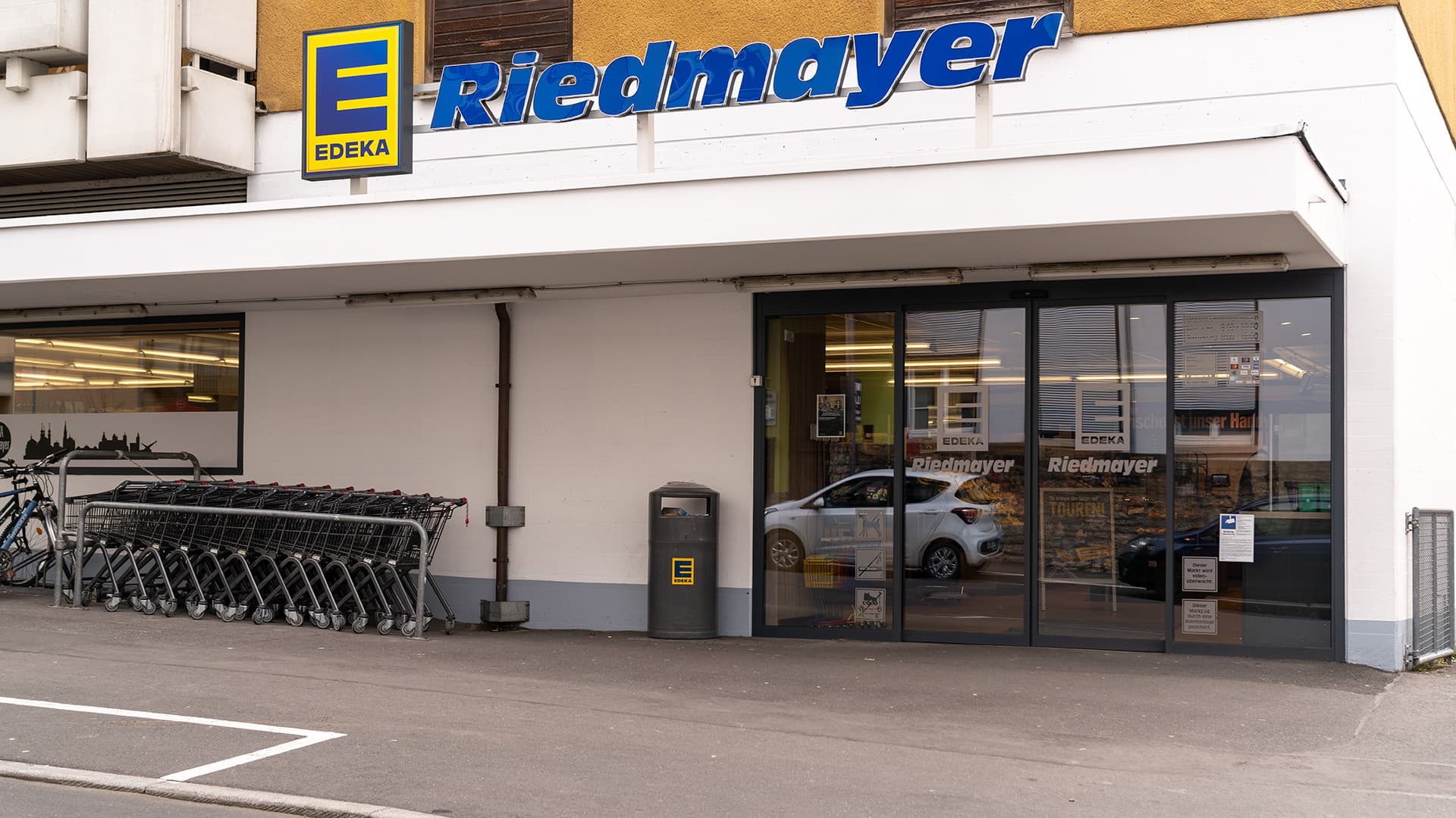 Eingangstür von EDEKA Riedmayer. Davor ein Mülleimer und Einkaufswagen.