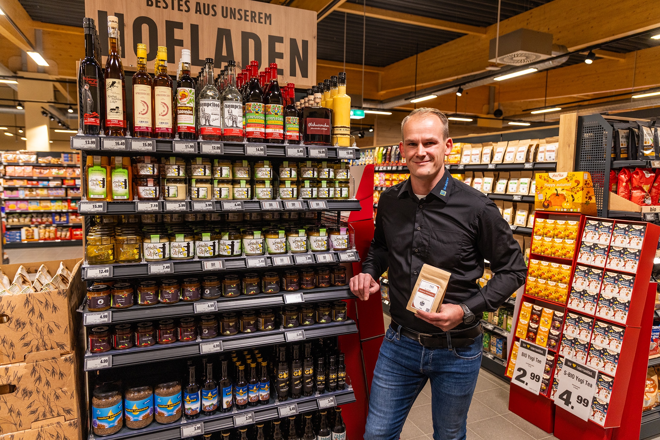 Marktinhaber steht vor dem Hofladenregal mit den regionalen Produkten und hält ein Produkt in der Hand.