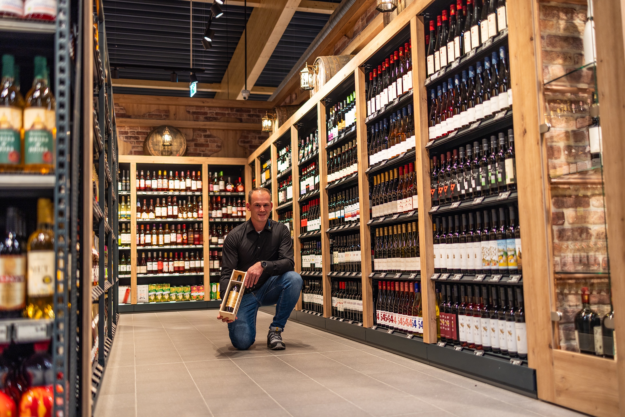 Der Marktinhaber im Gang der Weinabteilung kniend mit einer Weinflasche in der Hand.