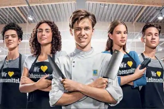 Fünf junge Menschen stehen nebeneinander und tragen Schürzen mit der Aufschrift: "Wir lieben Lebensmittel". In der Mitte steht eine Person mit verschränkten Armen und jeweils einem Messer in jeder Hand.