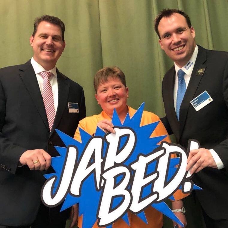 Drei Personen lächeln und halten gemeinsam ein Schild mit der Aufschrift: "Jap Bed!" in den Händen.