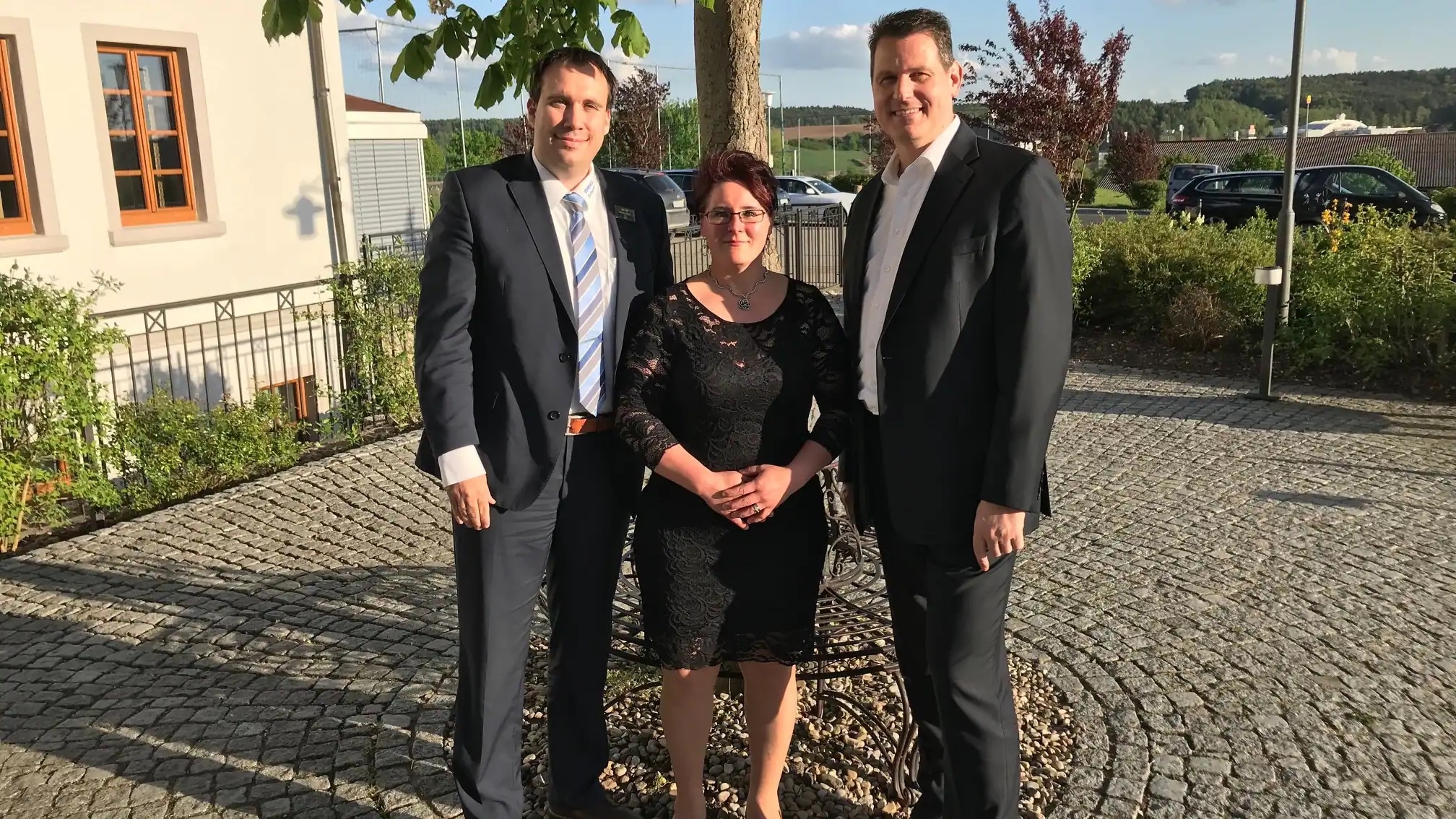 Zwei Männer im Anzug und eine Frau im Kleid stehen vor einem Baum und lächeln.