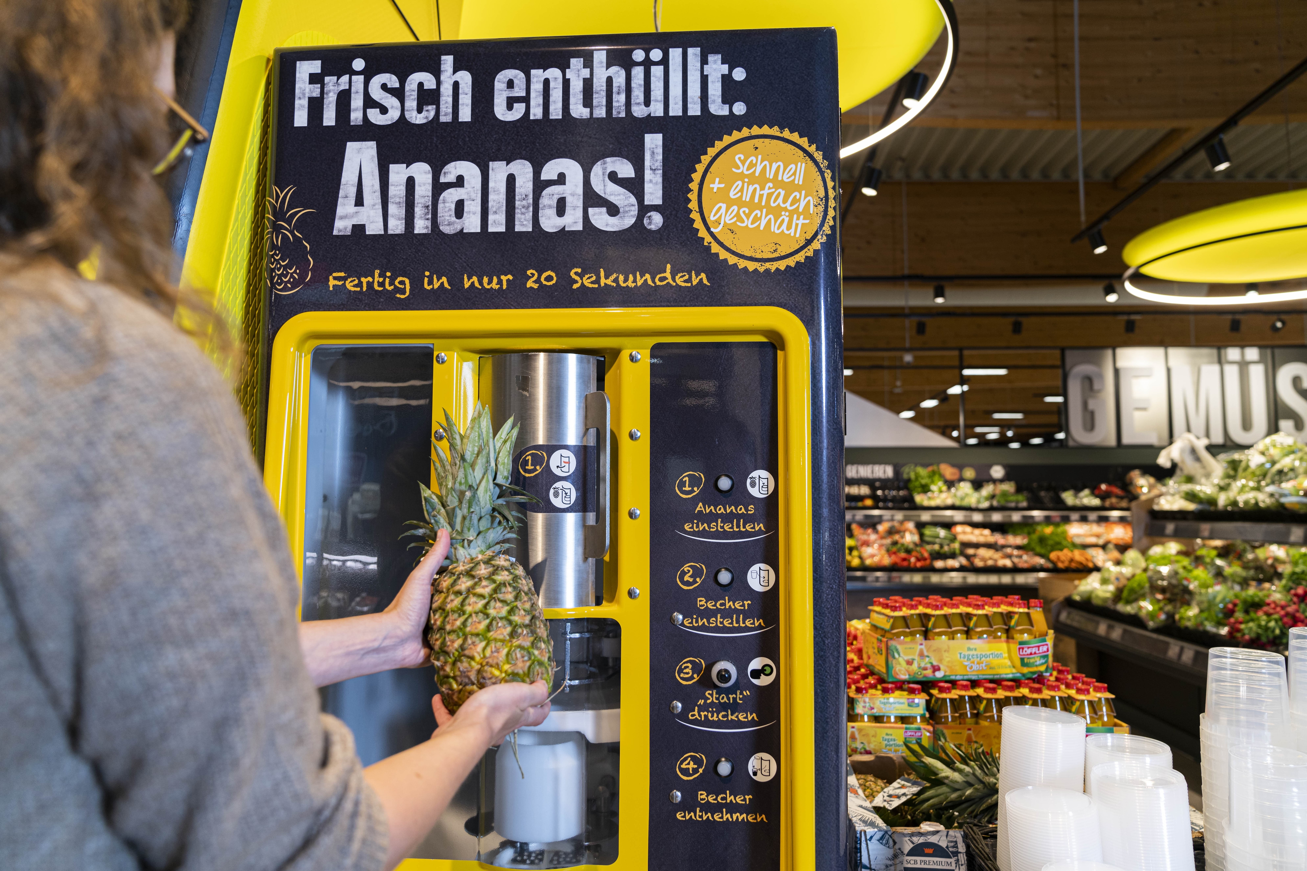 Eine Frau steht vor einem Automaten, auf dem steht: Frisch enthüllt: Ananas!
