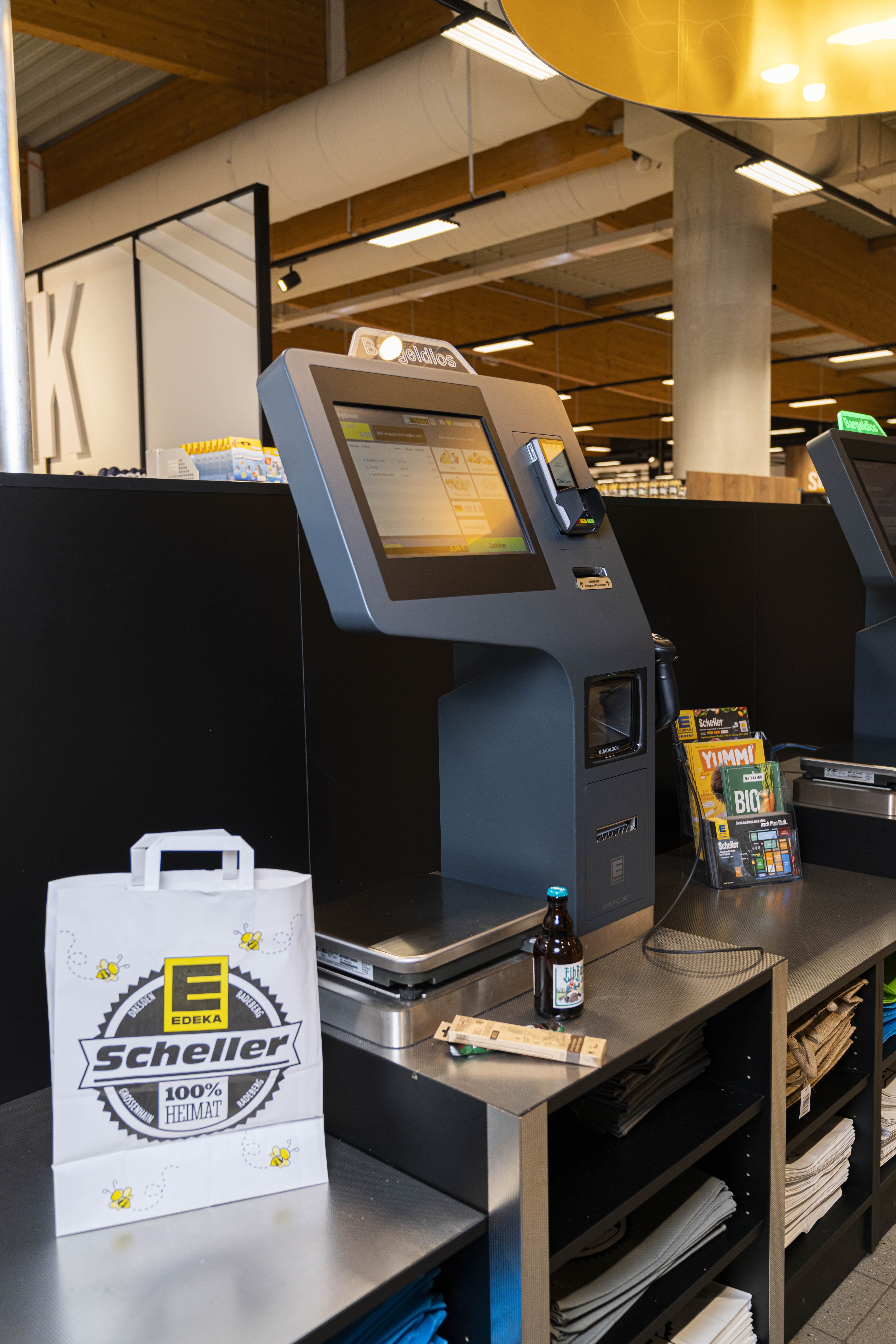 Selbstbedienungskasse mit Waage. Daneben steht eine Einkaufstüte aus Papier mit EDEKA Scheller-Logo.