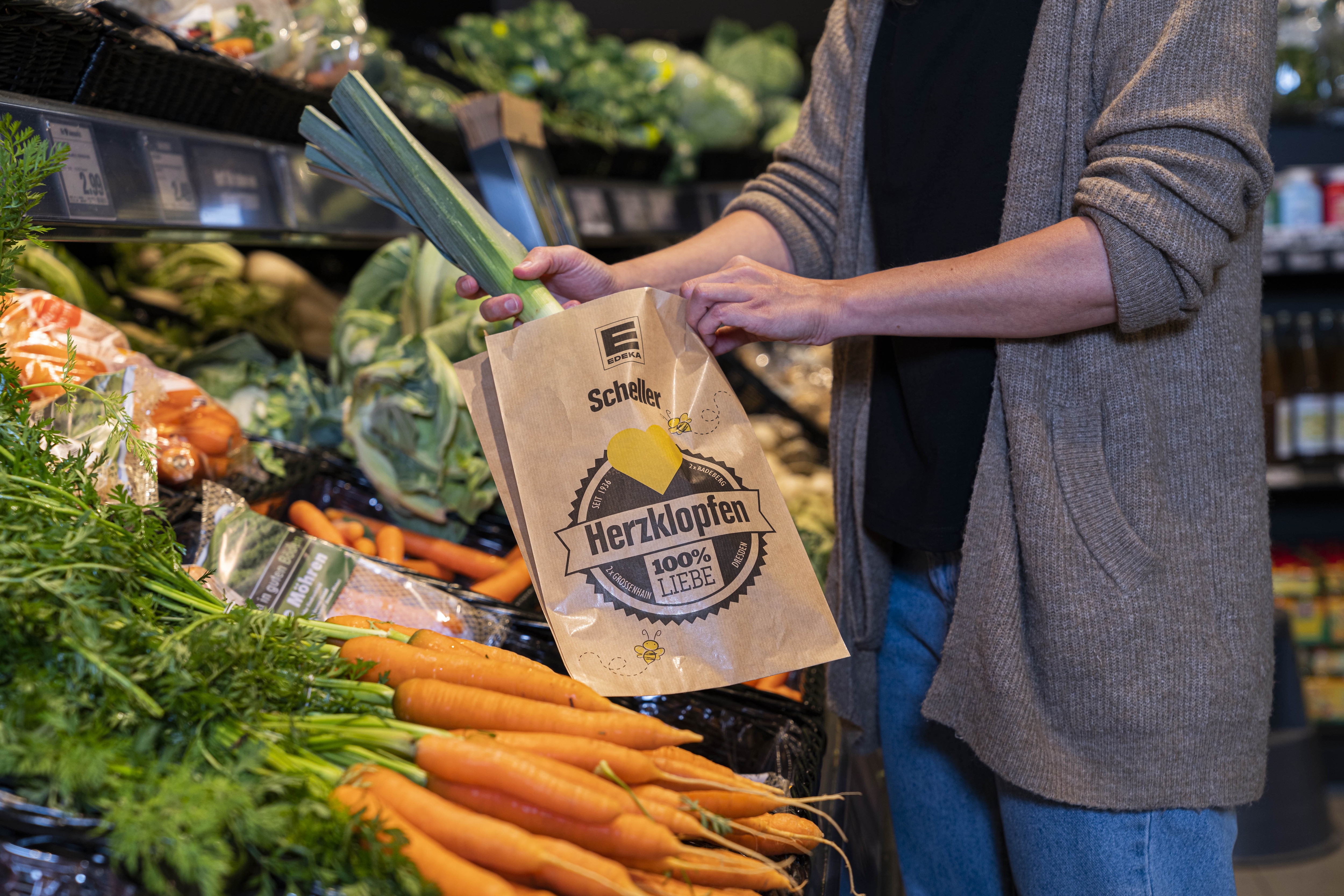 Eine Person mit Strickjacke legt einen Lauch in eine braune Papiertüte.