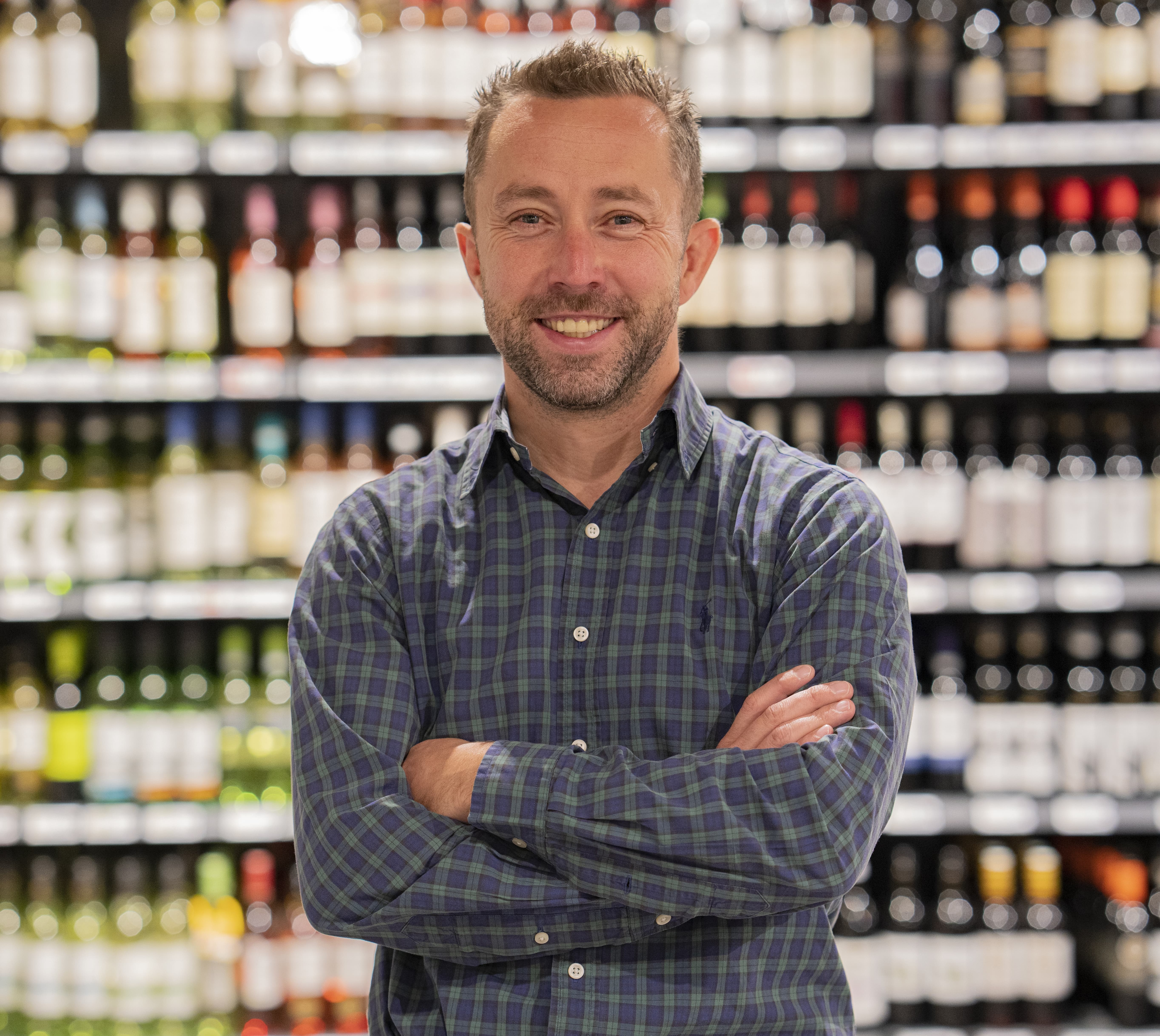 Ein Mann mit Dreitagebart steht grinsend vor einem Weinregal. Er trägt ein kariertes Hemd, seine Arme sind vor dem Oberkörper verschränkt.