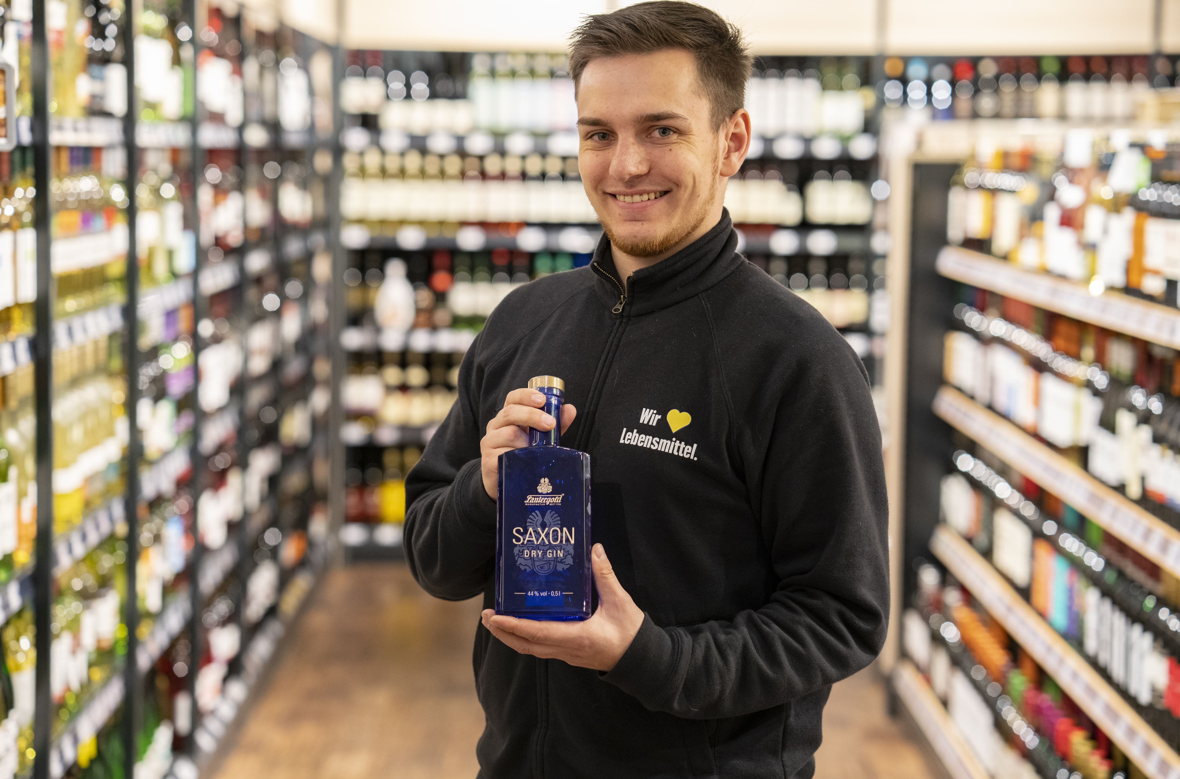Ein Mann in schwarzem EDEKA-Pullover hält eine blaue Flasche Saxon Gin hoch.