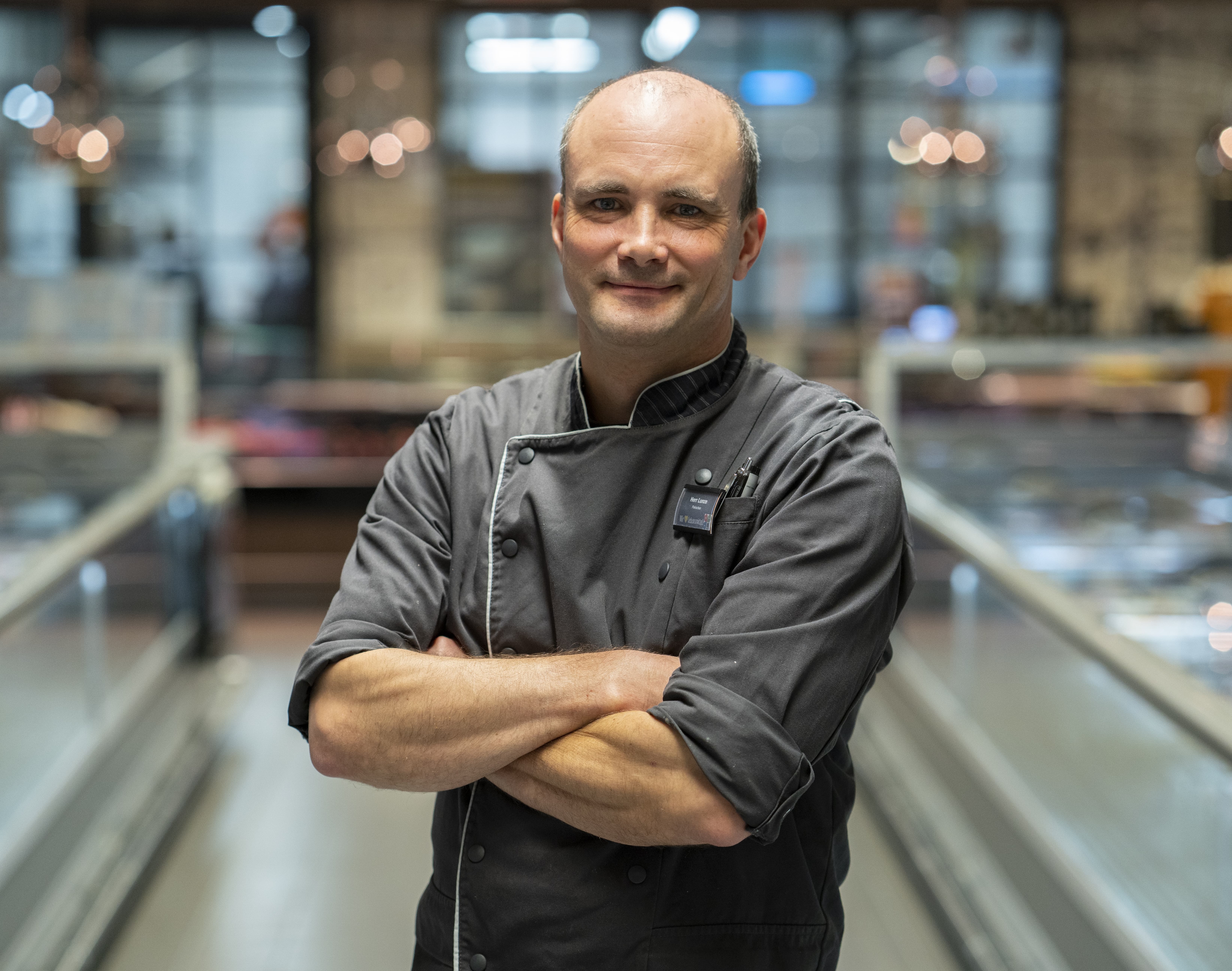 Ein Mann mit kurzen Haaren mit verschränkten Armen. Er trägt einen grauen Fleischerkittel.