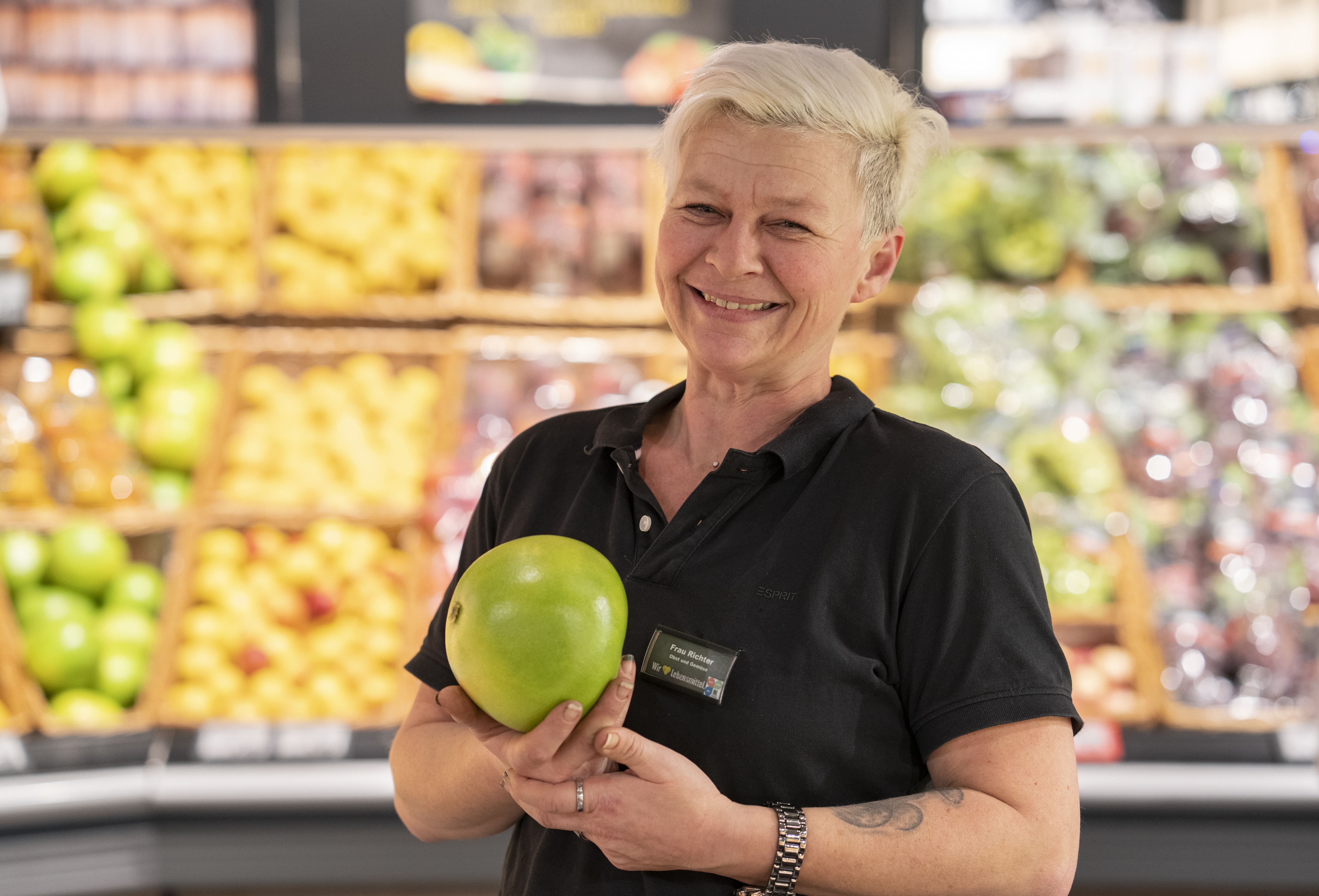 Eine Frau mit kurzen blonden Haaren und einem schwarzen EDEKA-Poloshirt hält eine große grüne Frucht hoch.
