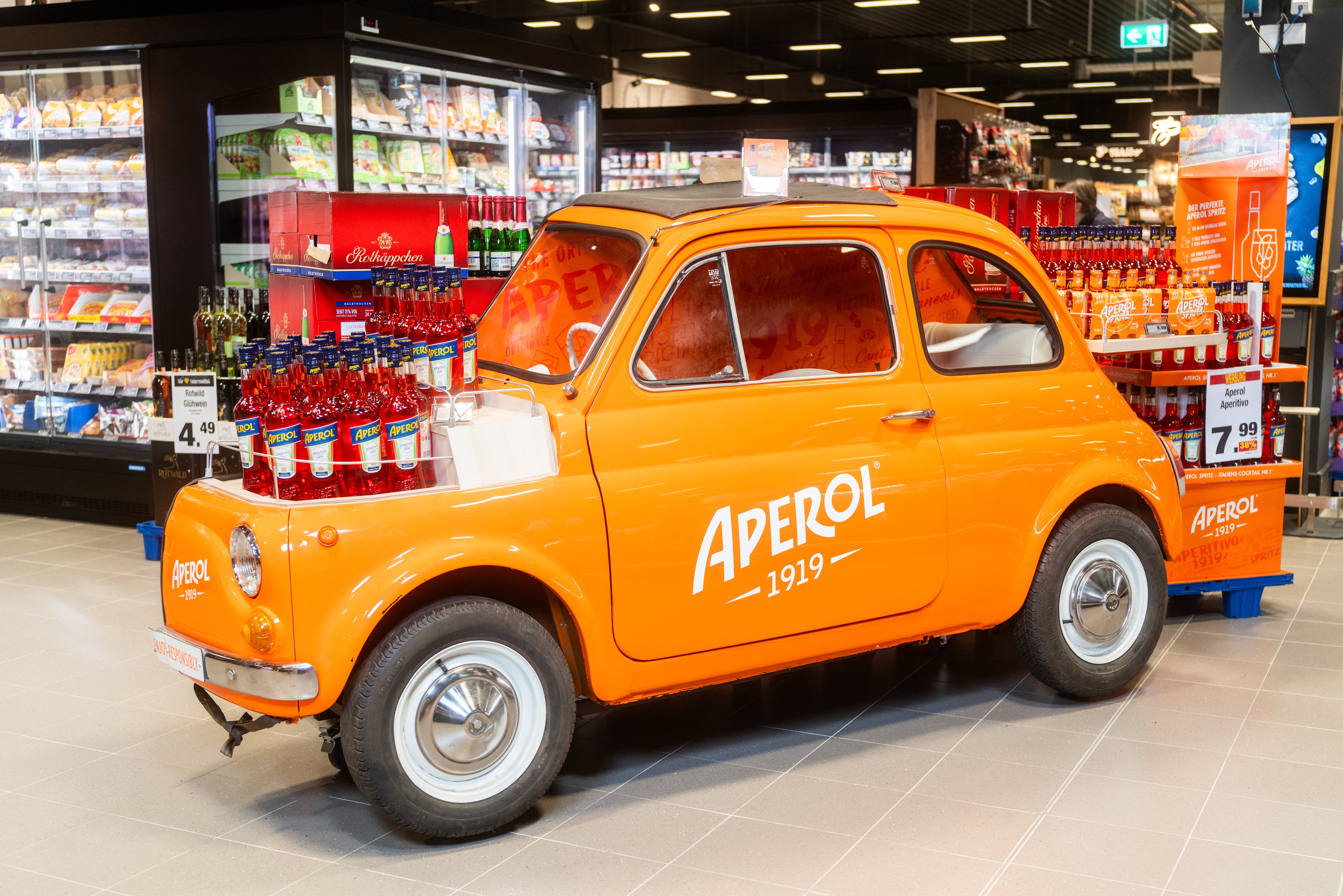 Ein orangener Kleinwagen mit der Aufschrift APEROL in einem Supermarkt. Auf der Kühlerhaube diverse Flaschen des roten Aperitifs.