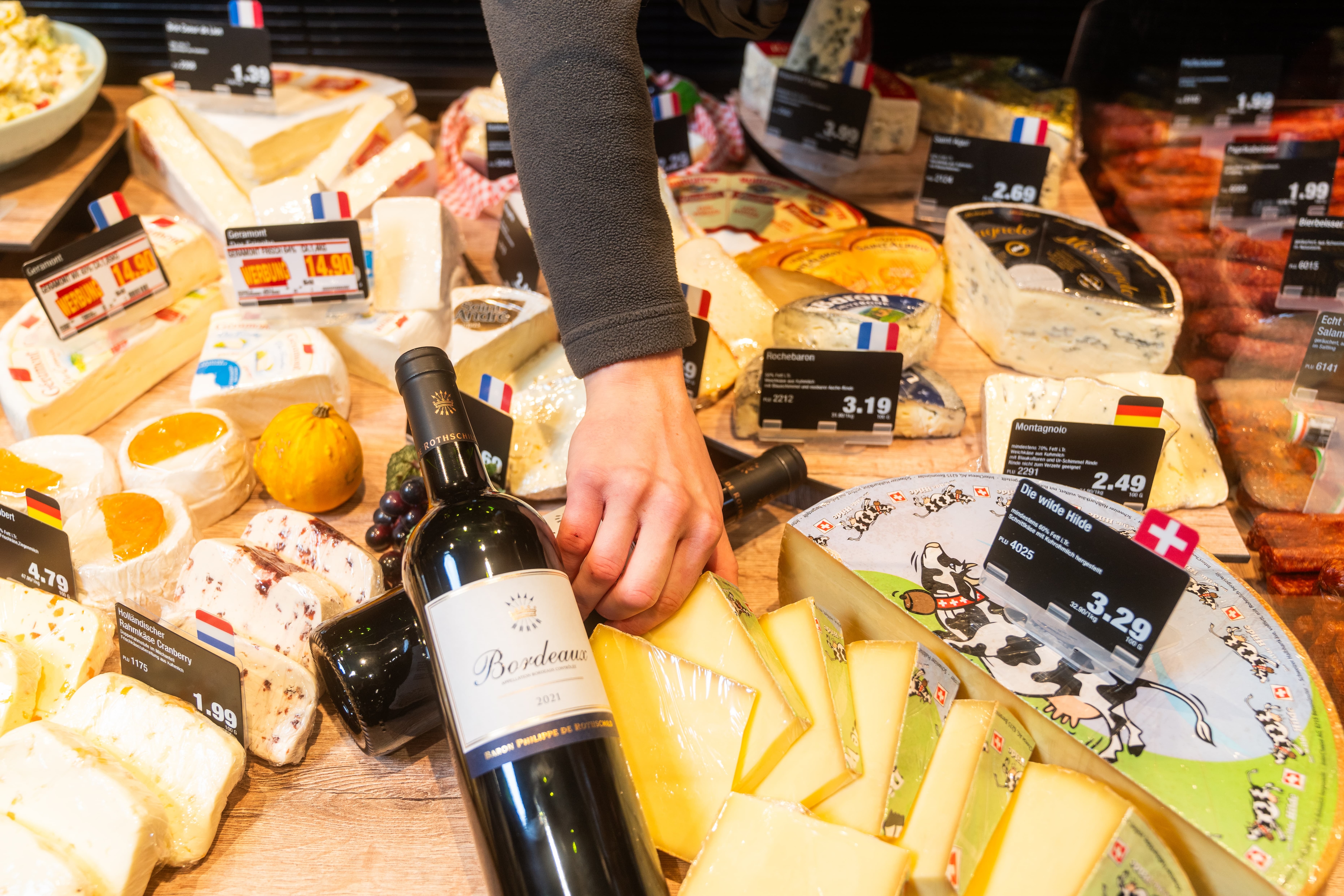 Verschiedene Käsesorten mit Preisschildern und eine Flasche Rotwein in einer Auslage.