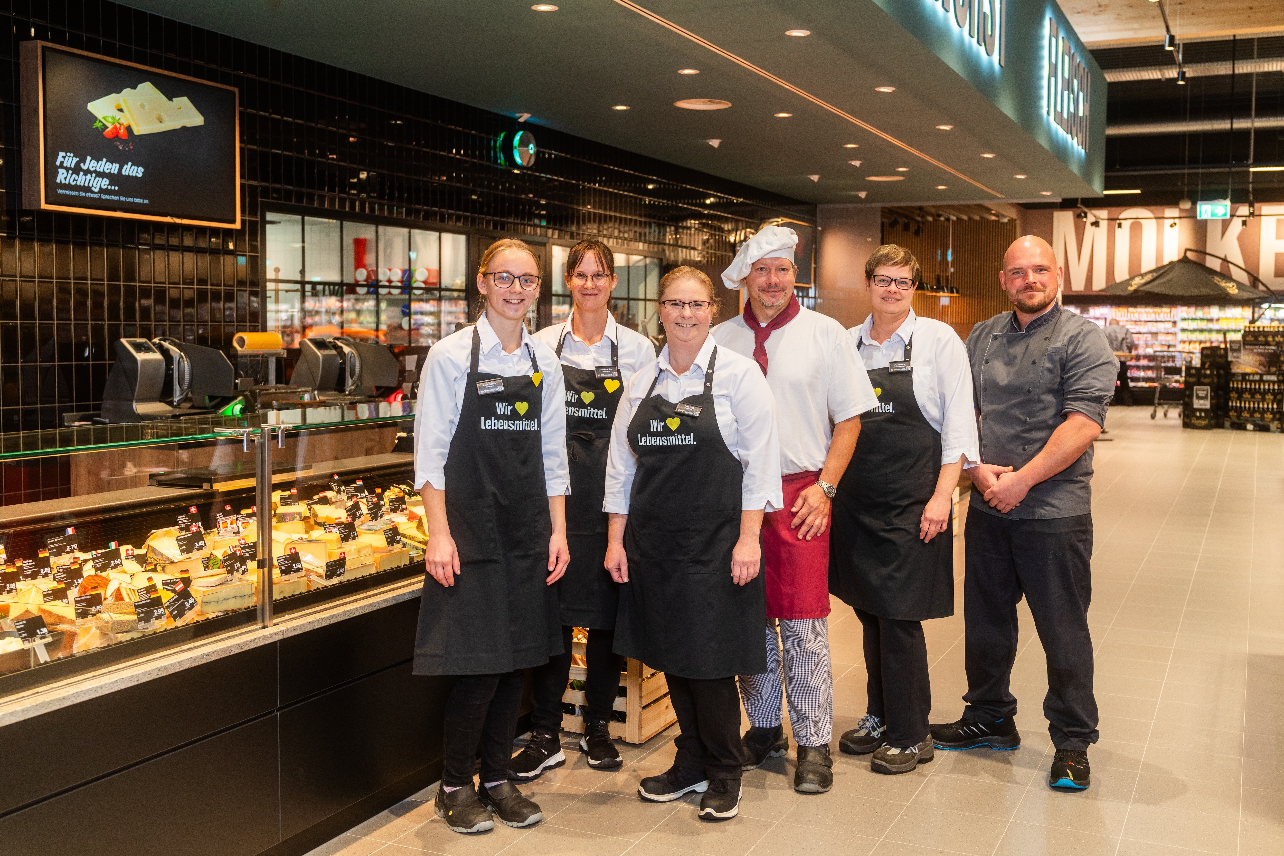 Vor der Frischetheke stehen sechs Personen. Vier von ihnen tragen schwarze EDEKA-Schürzen.