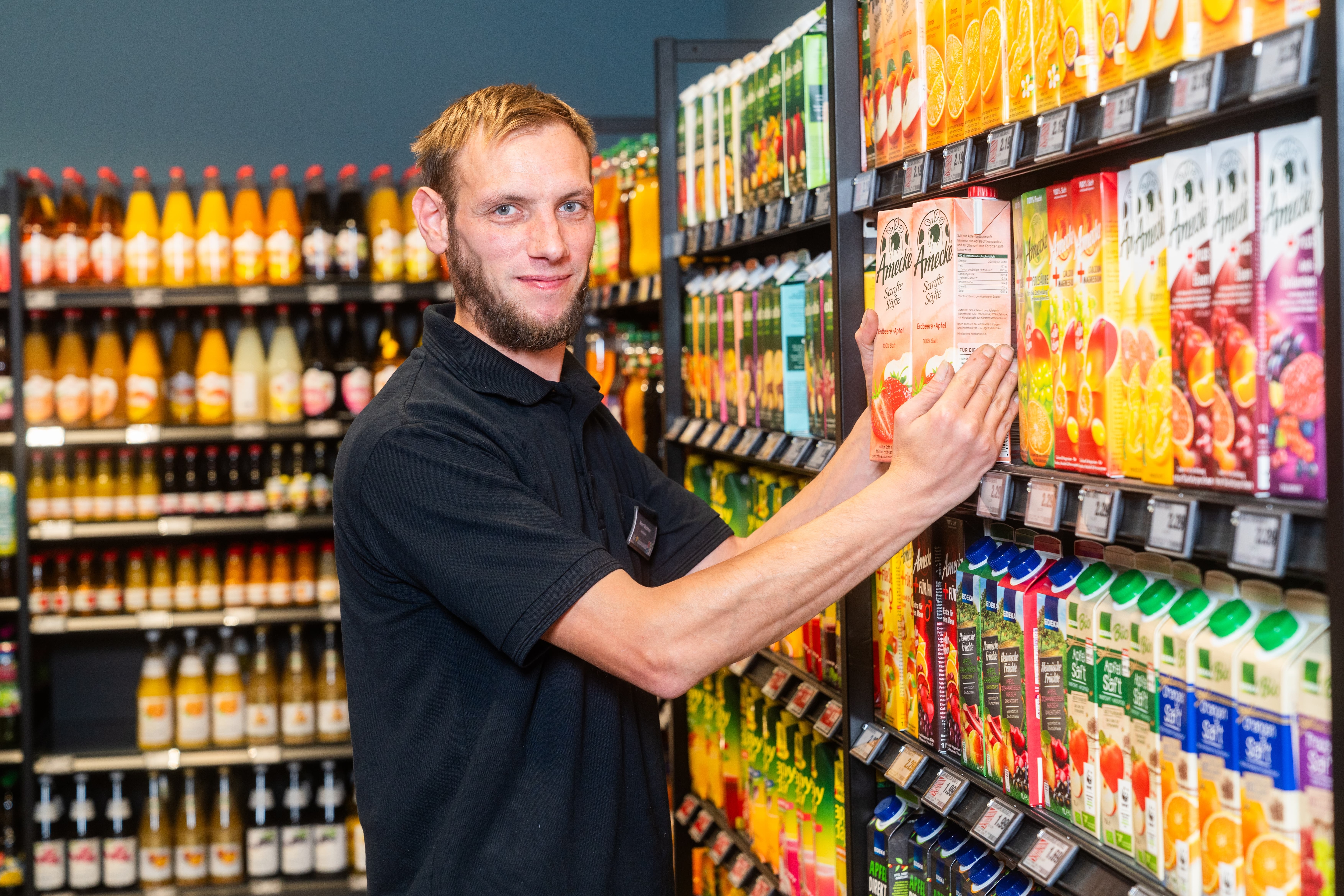 Ein junger Mann mit Bart nimmt zwei Tetrapacks aus dem Saftregal. Er trägt ein schwarzes Poloshirt.