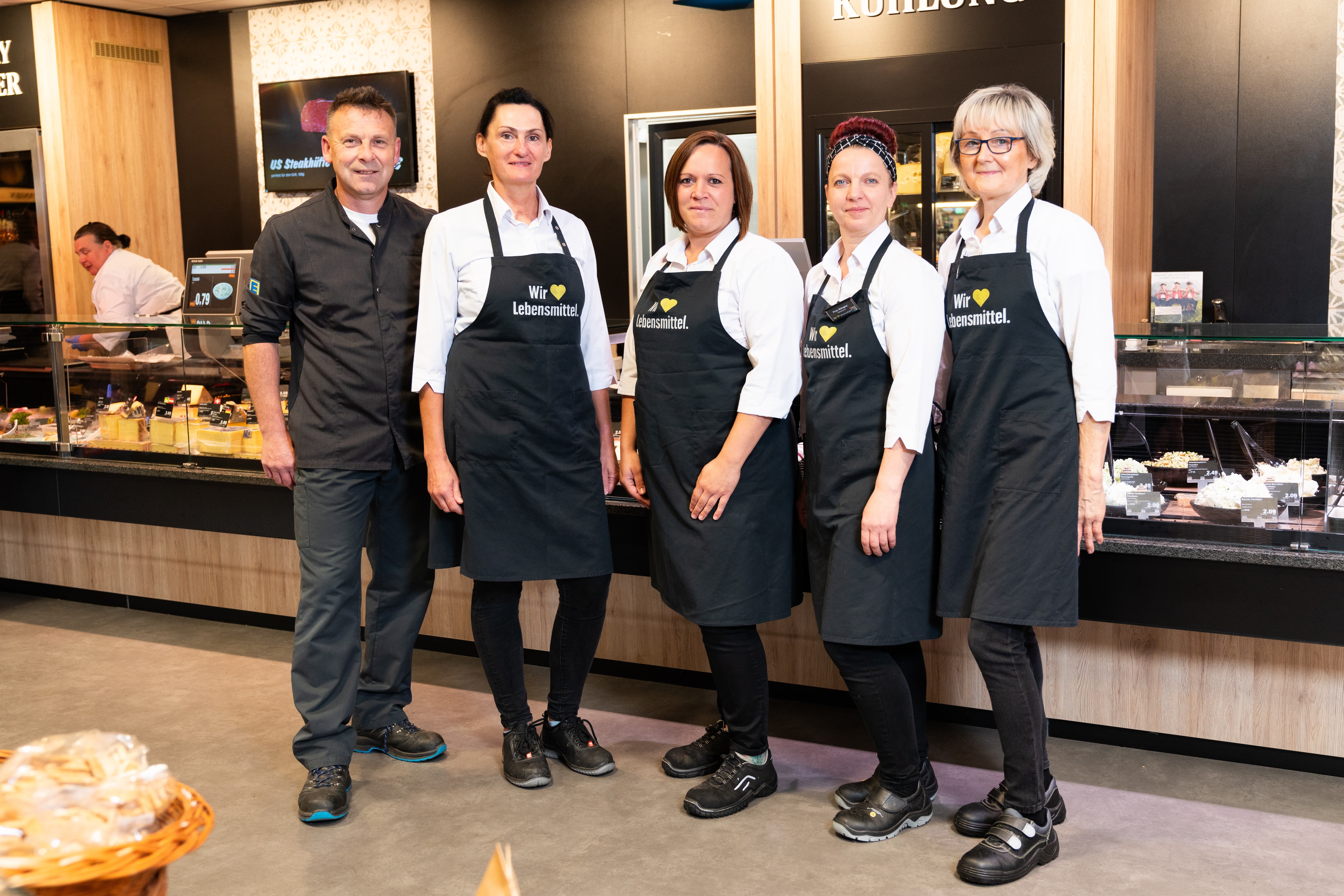 Vier Frauen in schwarzen EDEKA-Schürzen sowie ein Mann in schwarzem EDEKA-Hemd stehen vor der Frischetheke.
