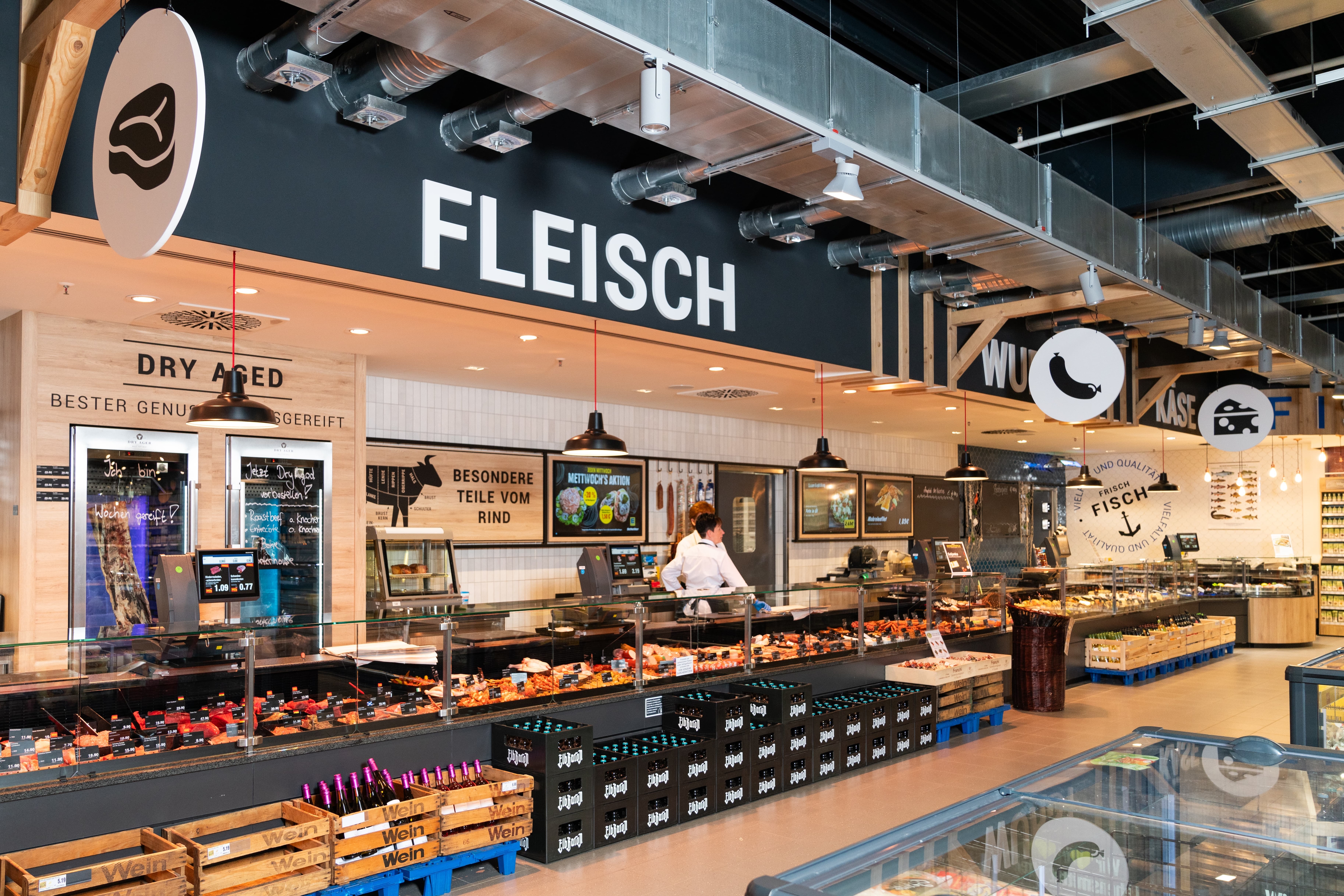 Hinter der Frischetheke stehen zwei Mitarbeitende in weißen Kitteln. Vor der Theke stehen Bierkisten.