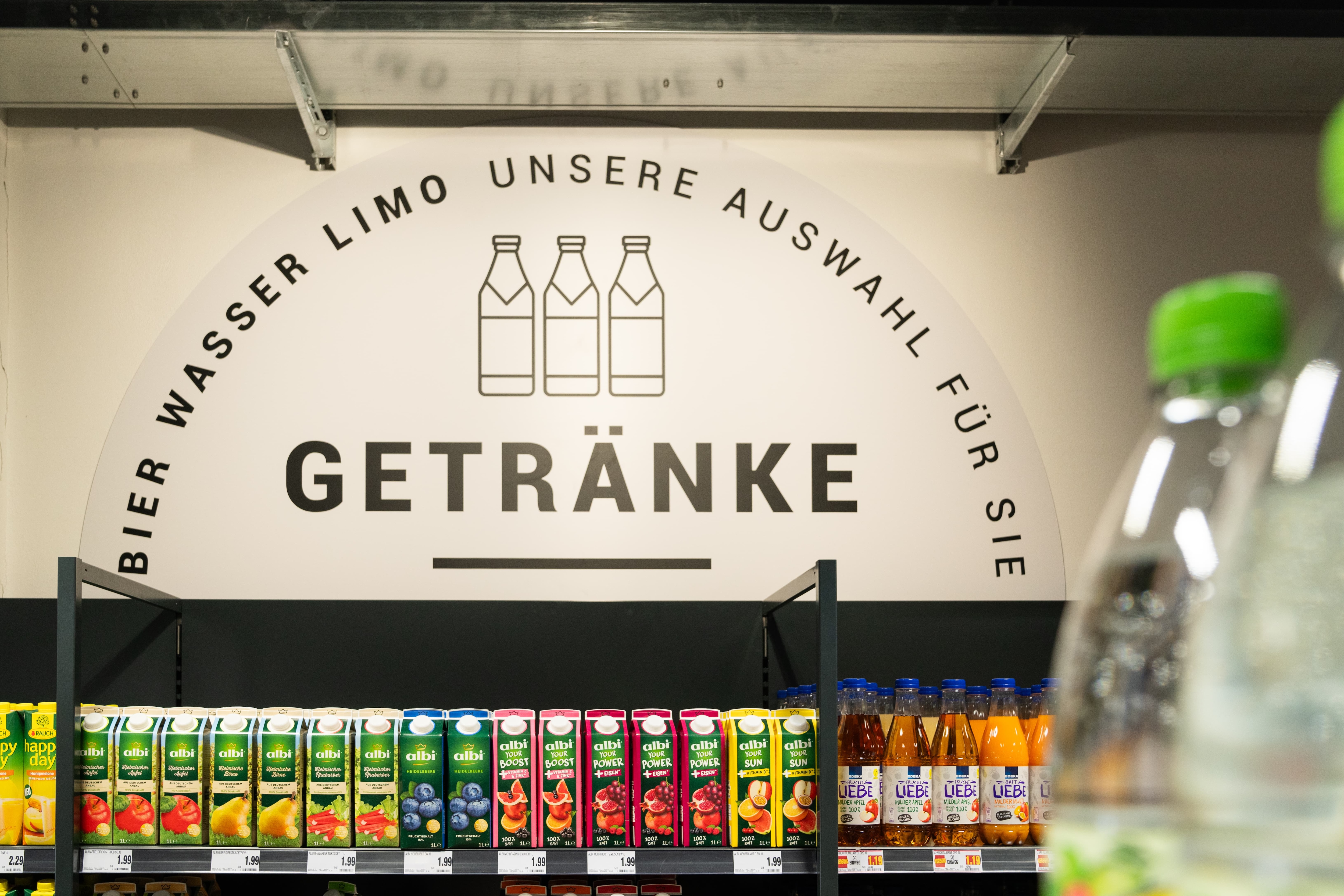 Über dem Saftregal hängt eine Illustration mit drei Flaschen und dem Wort Getränke.