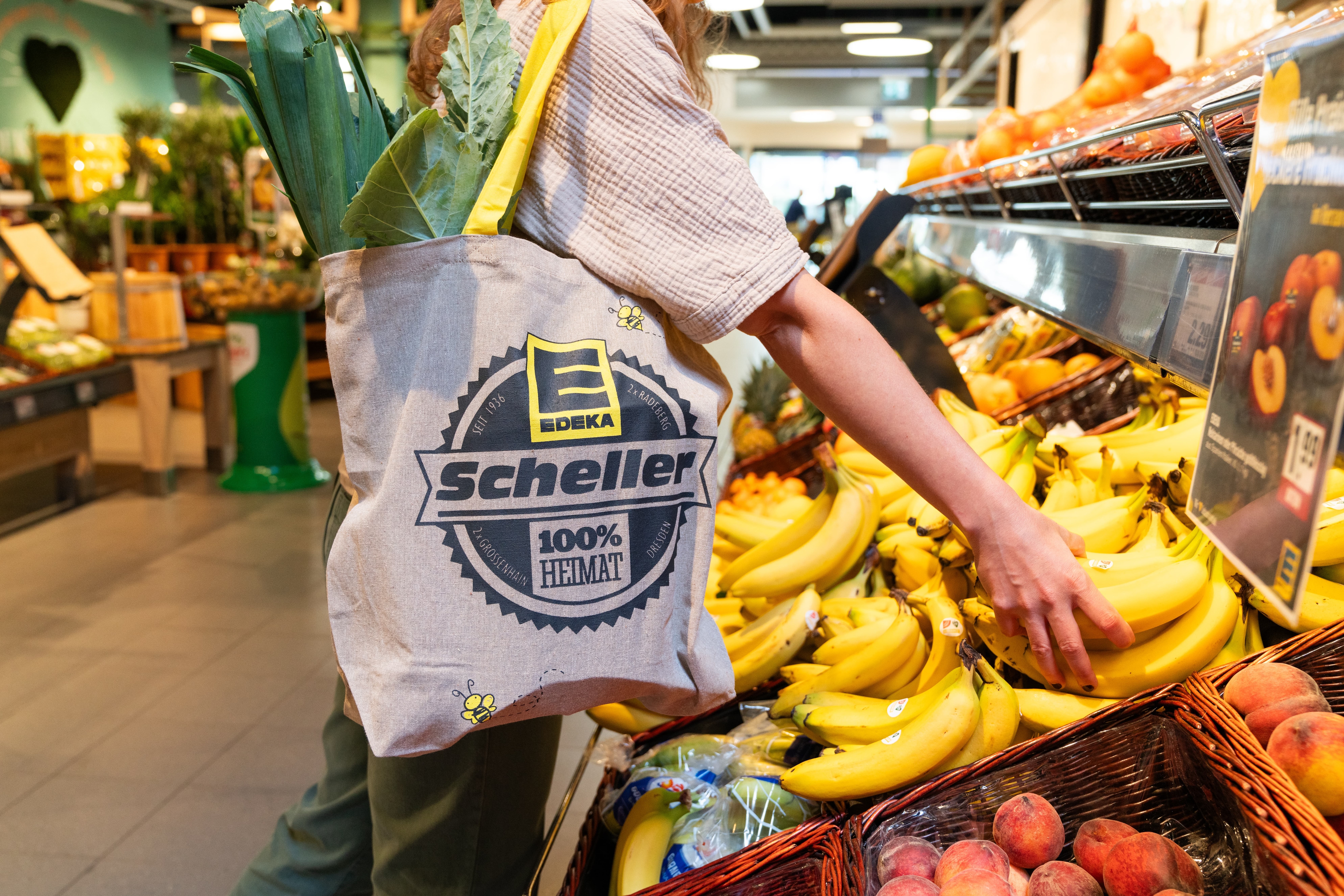 Eine Person greift nach Bananen aus dem Obstregal. Über ihrer Schulter hängt ein Tragebeutel mit dem Logo von EDEKA Scheller.