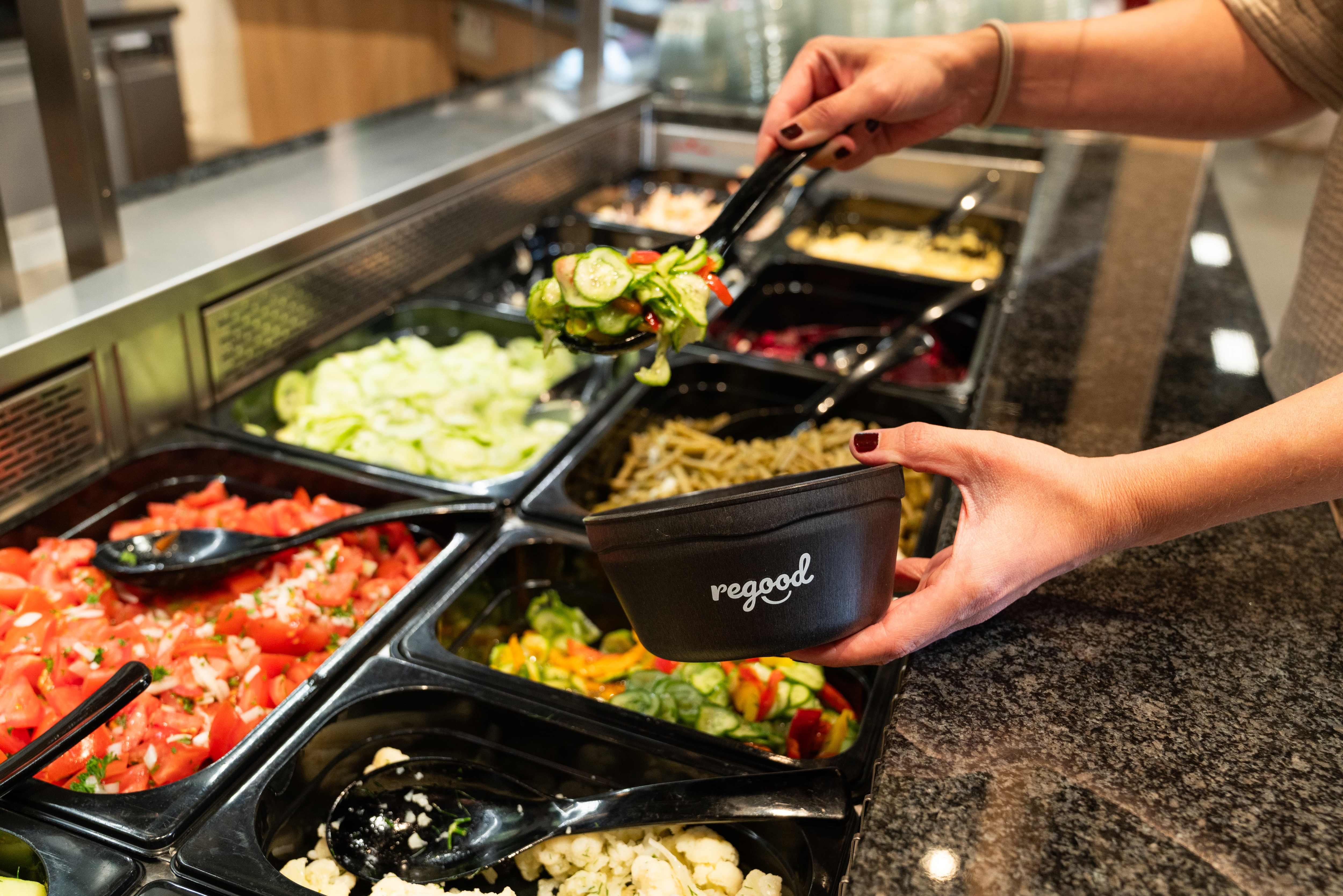Eine Person füllt Gurkensalat in einen schwarzen Mehrweg-Behälter der Firma regood.