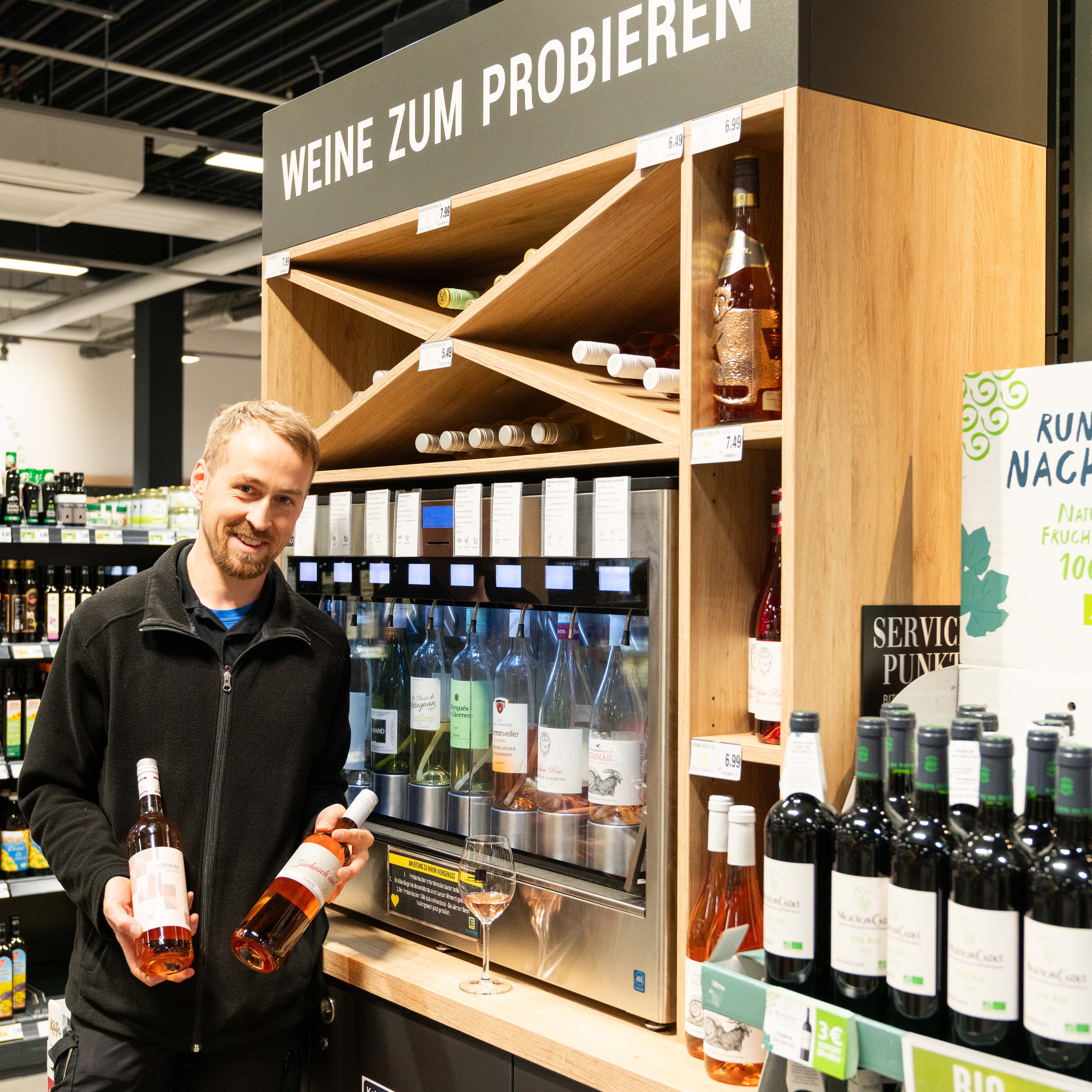 Ein Mann steht vor einem Zapfregal für Wein und hält jeweils eine Flasche in jeder Hand.
